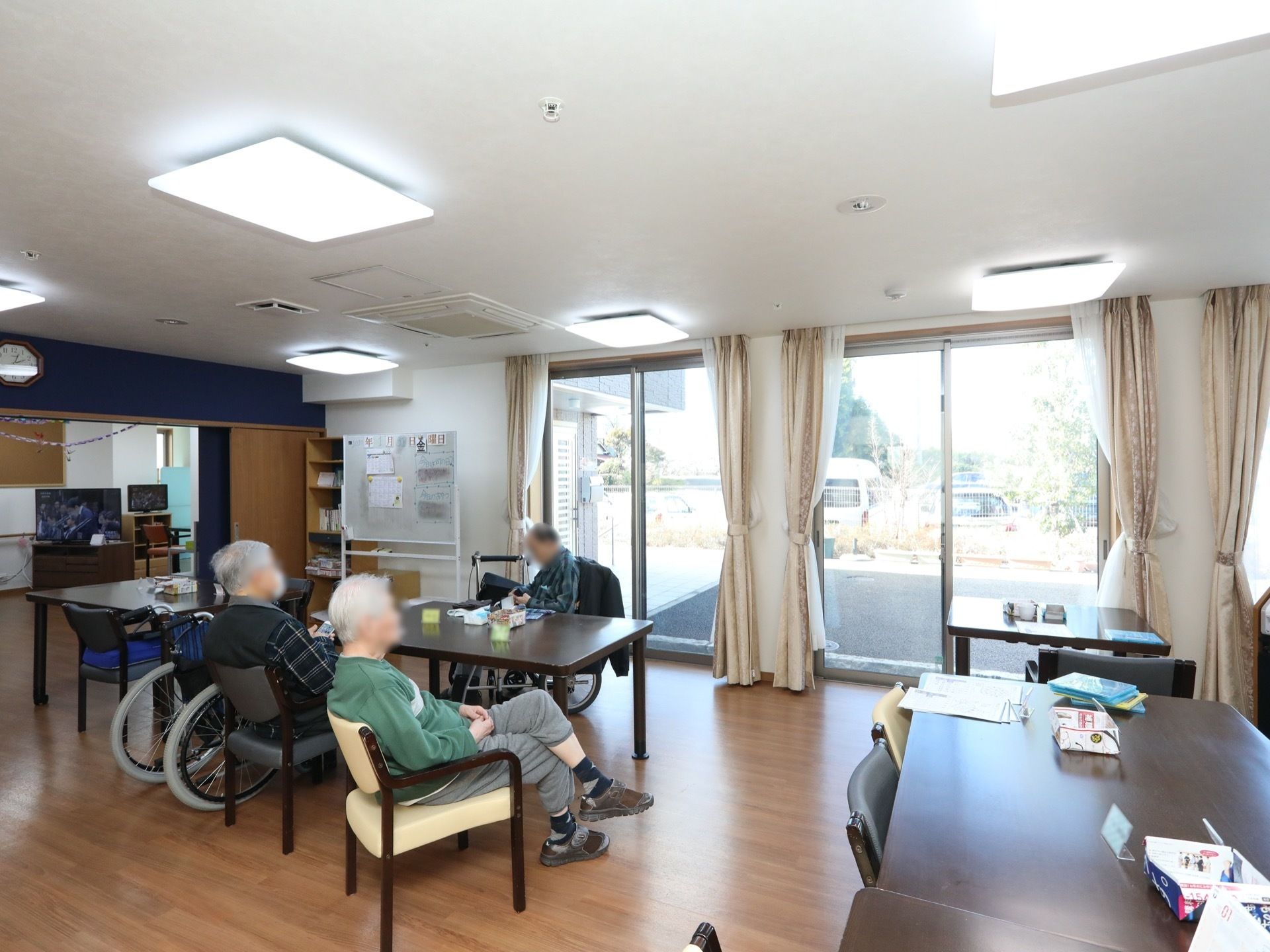 男性の入居者様が3人、テーブルを囲んで座っている。壁の近くには、ホワイトボードが置いてありプリントが貼ってある。