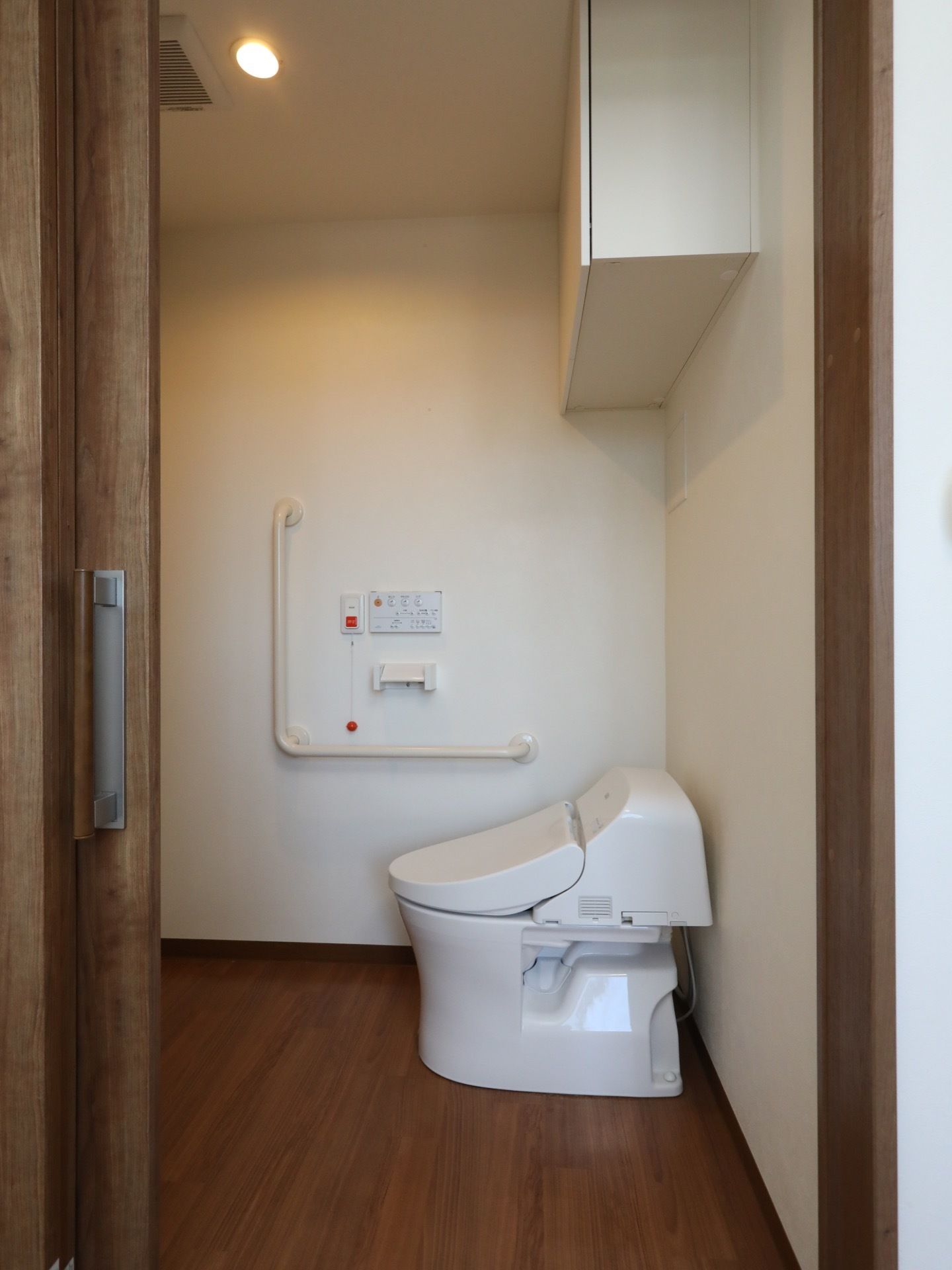 木製のドアに、大きなI字の手すりが付いたトイレである。壁には、L字の手すりや操作盤、緊急通報装置が取り付けられている。