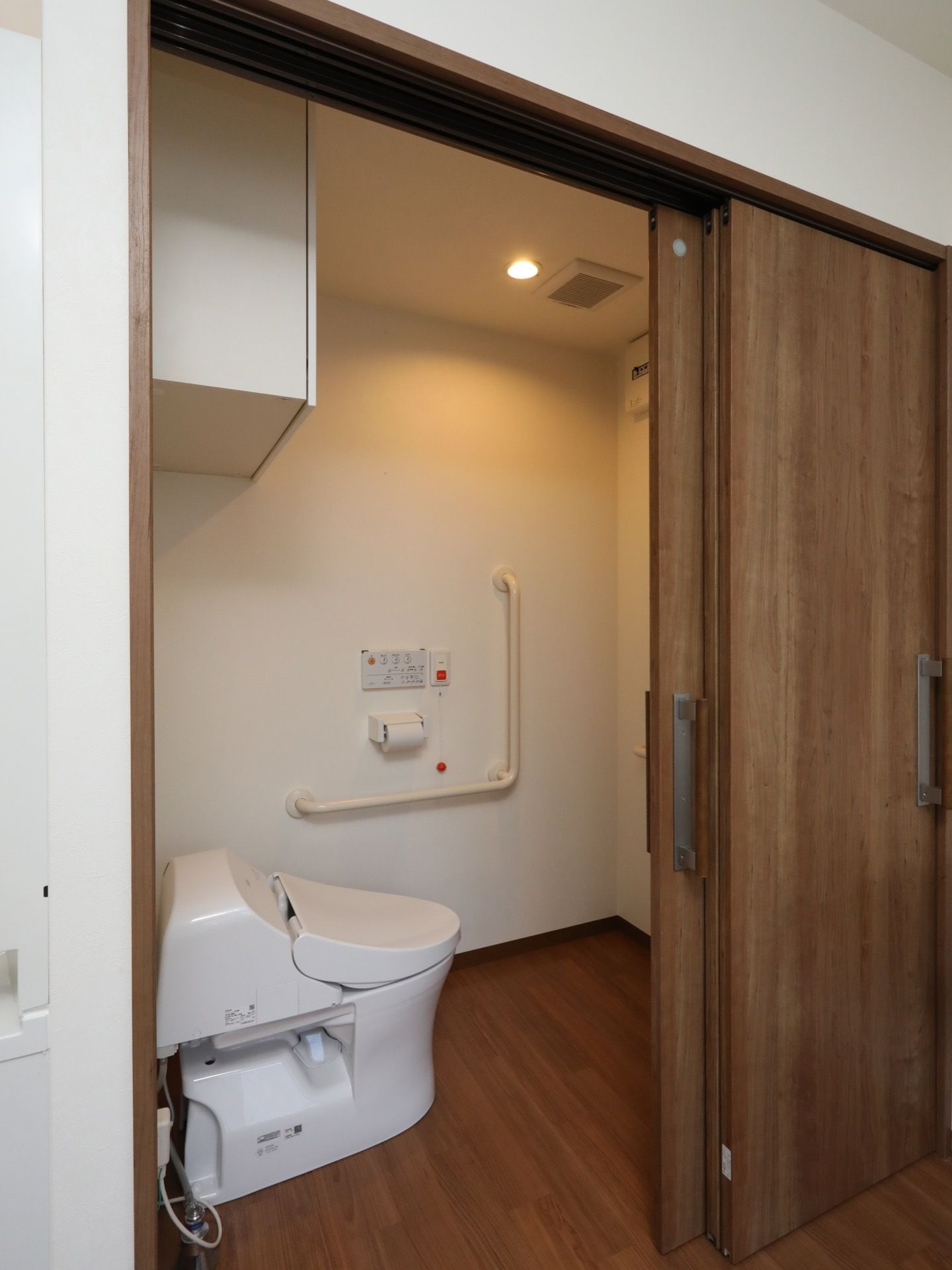 トイレのドアは、引き戸式となっている。温水洗浄機能付きの便座が設置されている。壁に、L字型の手すり、ナースコールなどが付いている。