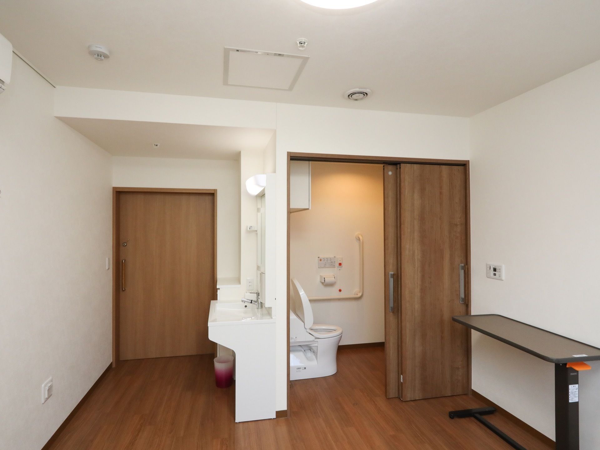 ドアのすぐ右側の所に洗面台があり、洗面台の後ろ側にトイレがある。トイレのドアの側には、オーバーテーブルが置いてある。