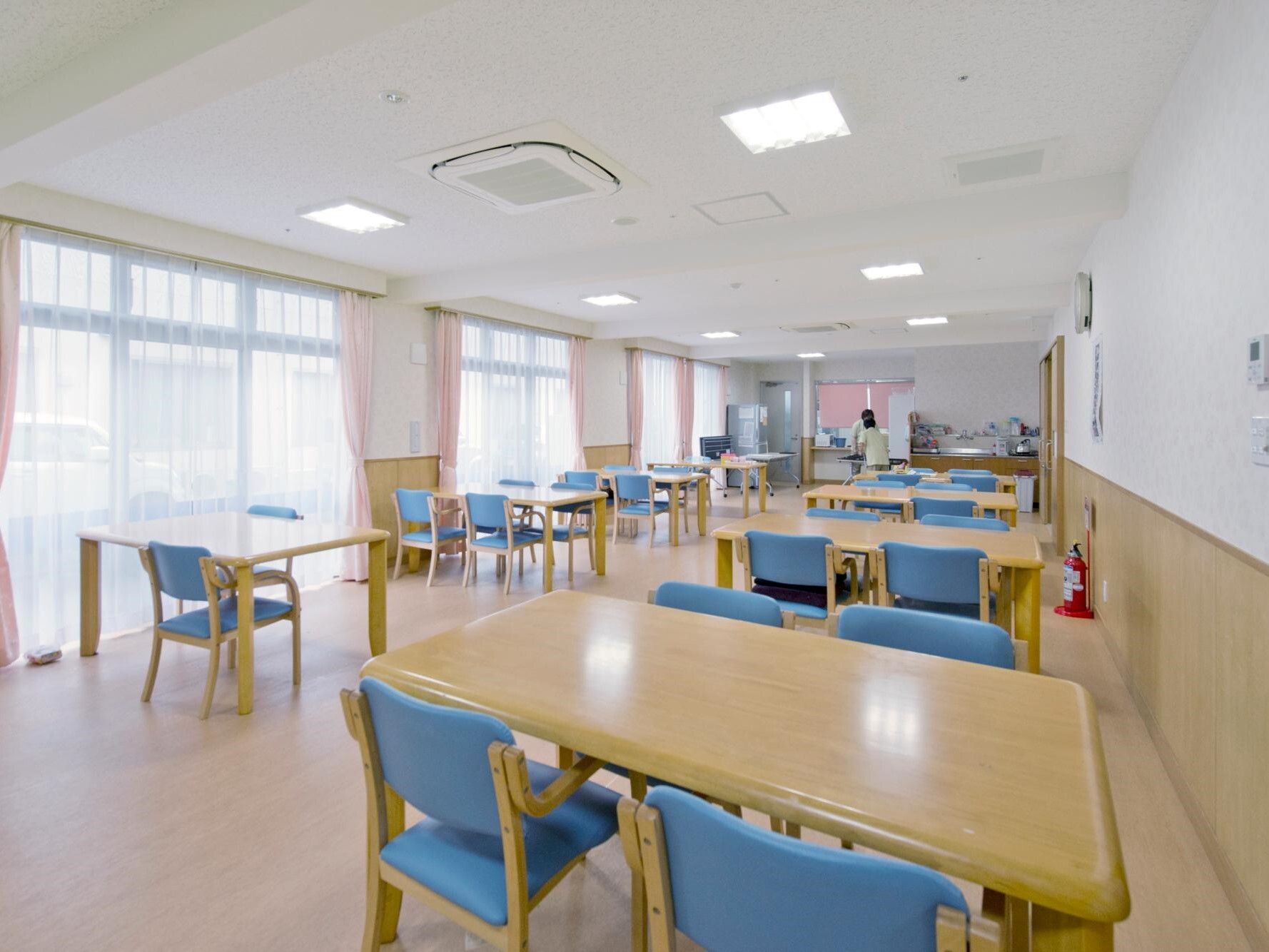 大きな窓があり明るい。ダイニングテーブルが複数置かれ、青い椅子がセットされている。