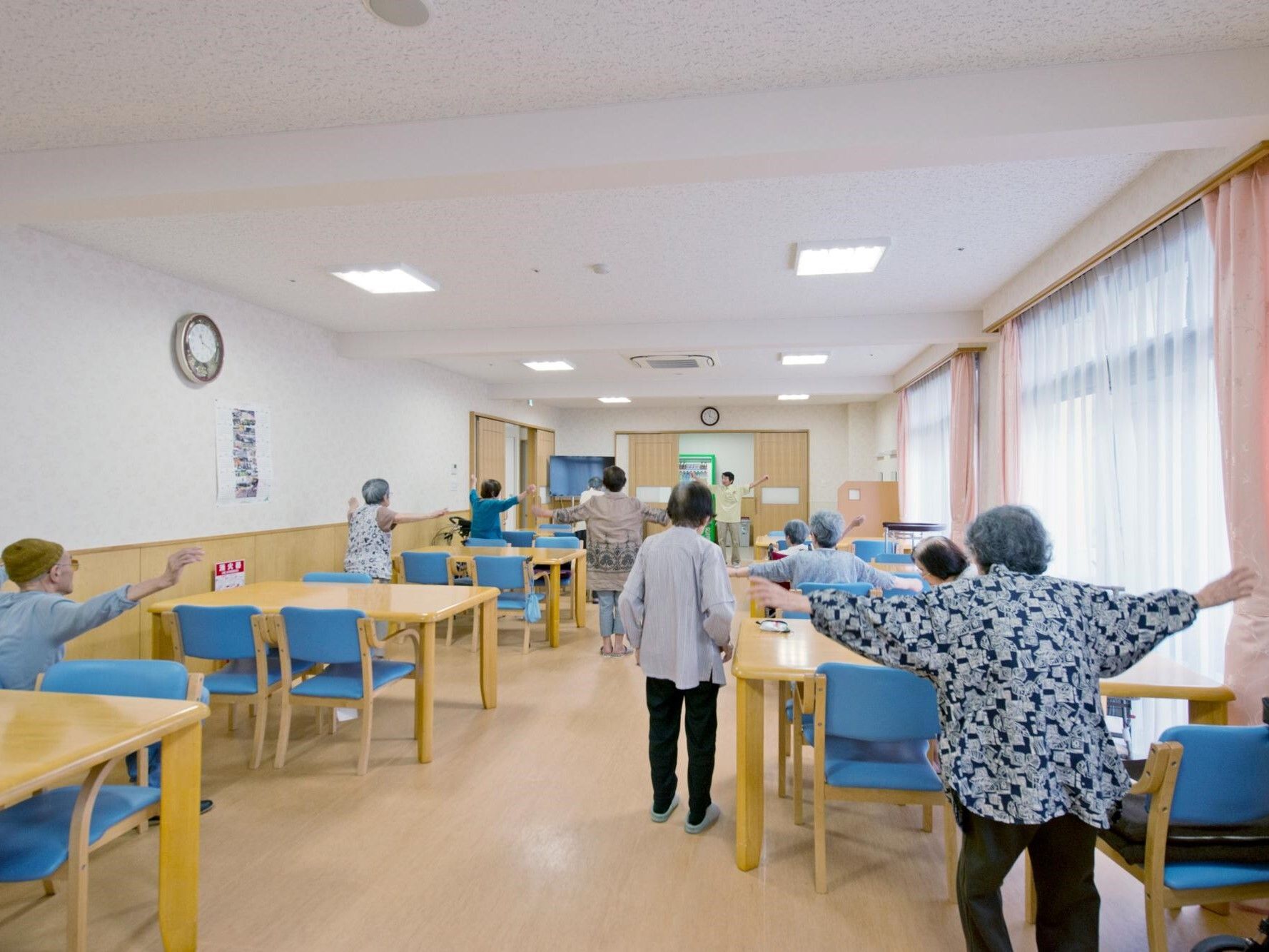 ダイニングテーブルが置かれた食堂で体操をしている入居者たち。前にスタッフが立ち、入居者は座っている人も建っている人もいる。