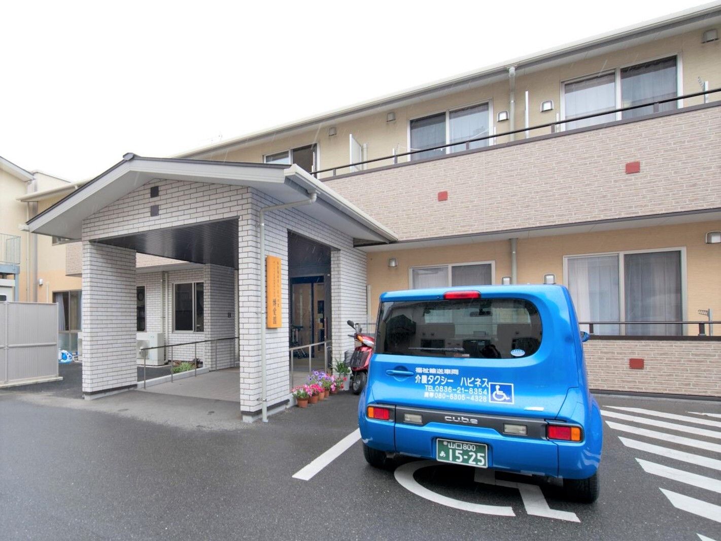 ベージュの2階建ての建物の玄関には屋根が付き、建物の前には駐車場も設置され、青い車が止められている。