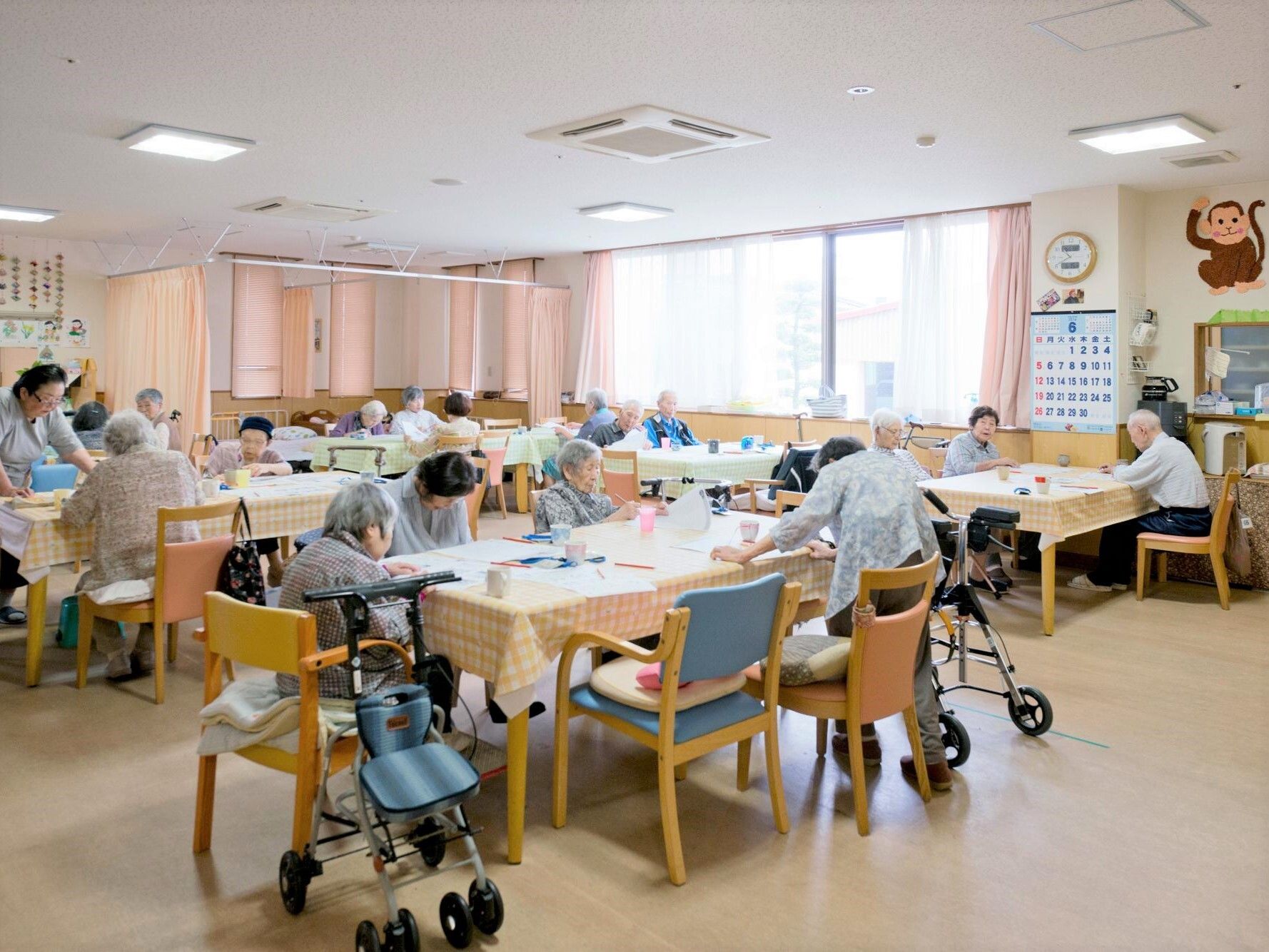 大きな窓があり明るい。ダイニングテーブルが複数置かれ、入居者がテーブルを囲んでいる様子。