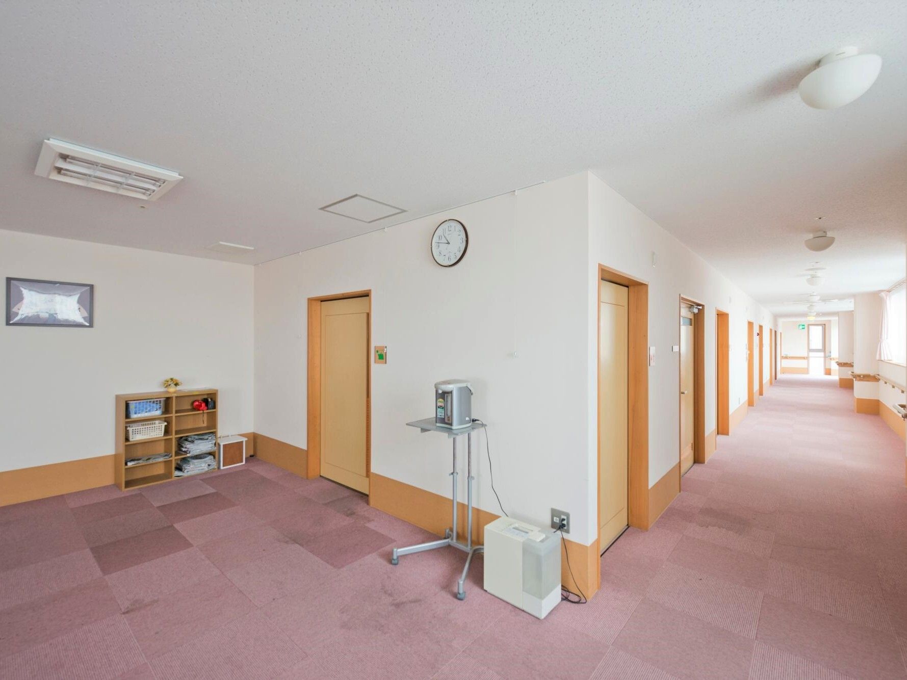 右側には居室のドアが並ぶまっすぐな廊下があり、左側には棚のようなものが置かれている。