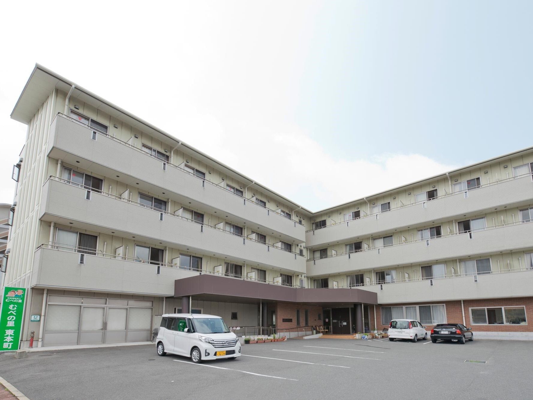 駐車場やベランダが付いた白を基調とした４階建ての建物。窓がたくさんつき、１階部分の出入り口には屋根が付いている。