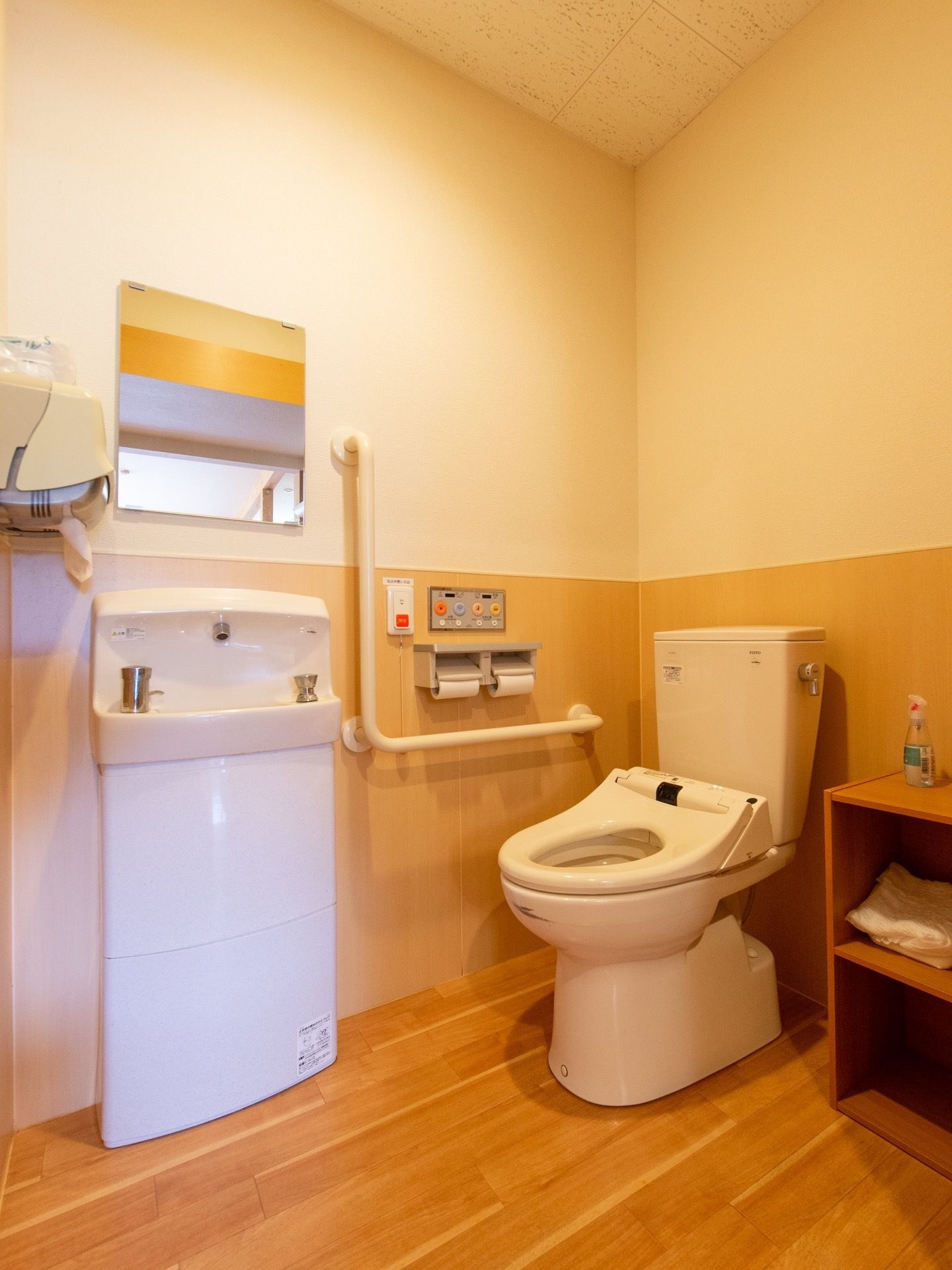 トイレの床はフローリングになっており、便器の横にはカラーボックスが置かれている。壁にはL型の手すりや呼び出しボタンなどが設置されている。