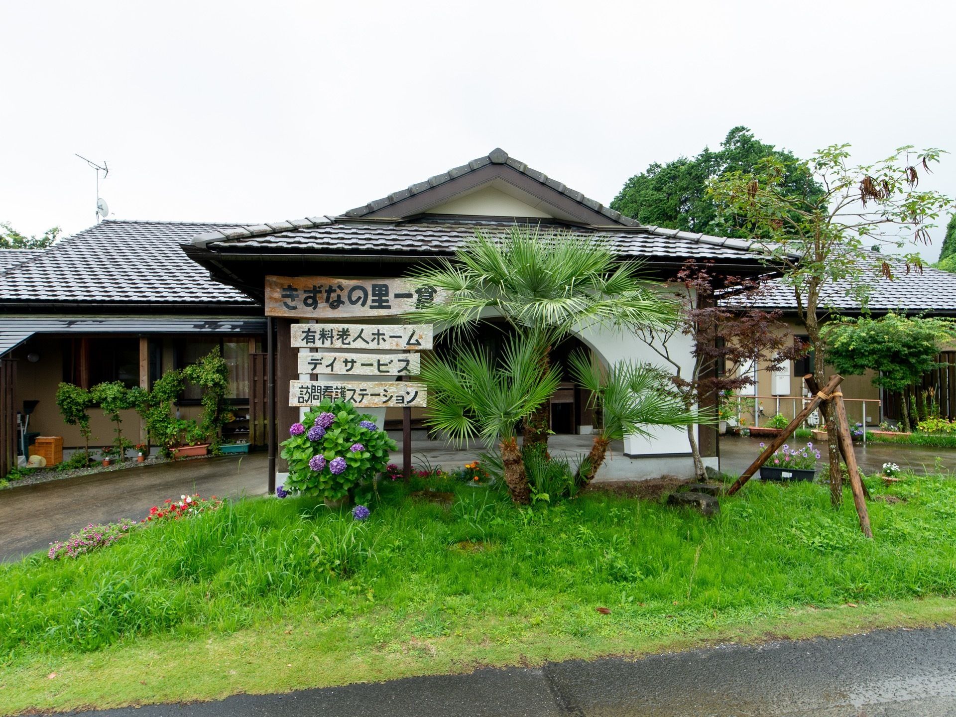 建物は瓦屋根の和風な造りである。玄関の前には木や花が植えられており、木製の施設の看板が付けられている。
