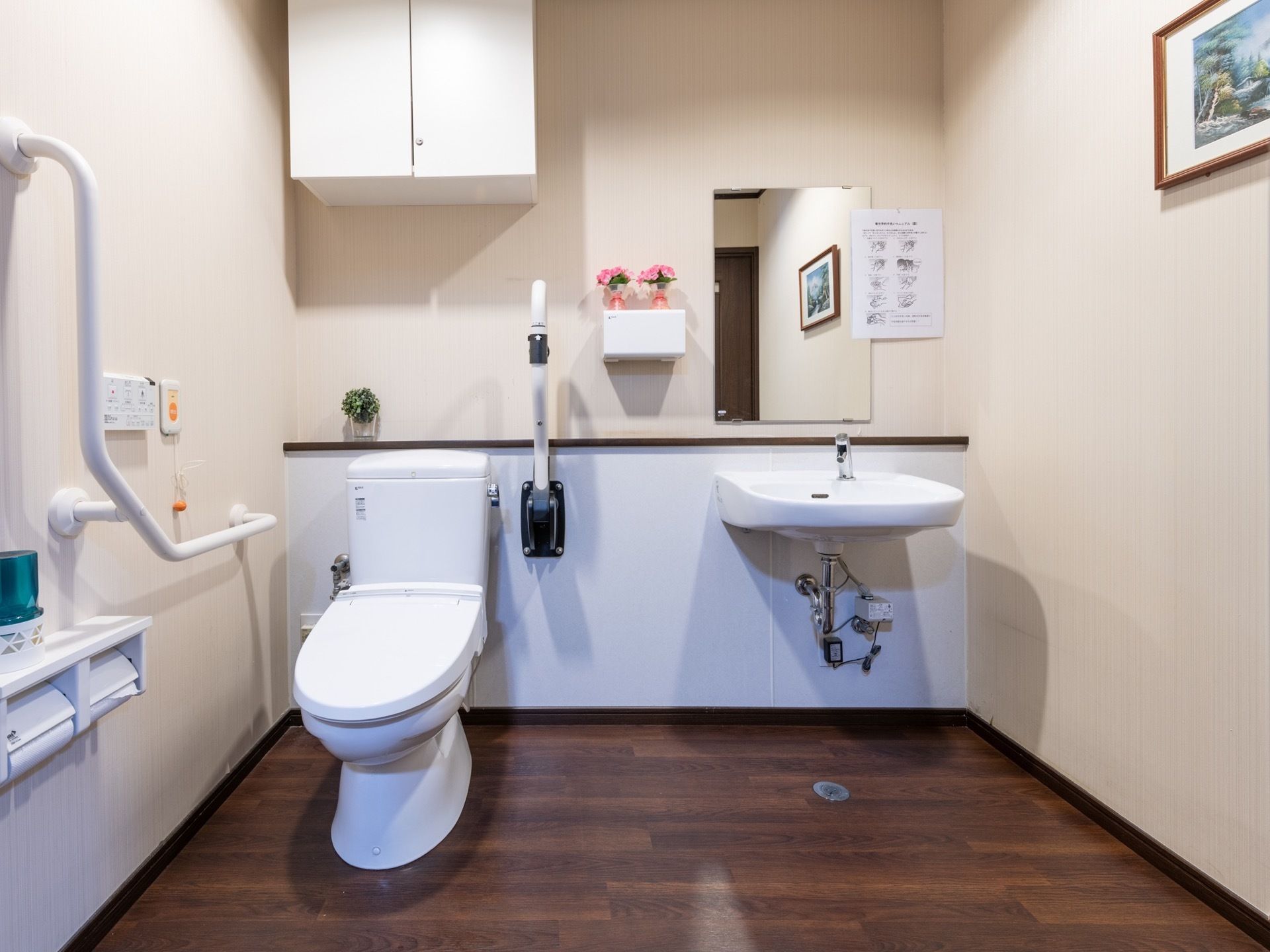 広く清潔感のあるトイレである。可動式の手すりや、緊急時呼び出しボタンが備わっており、数カ所に花が飾られている。