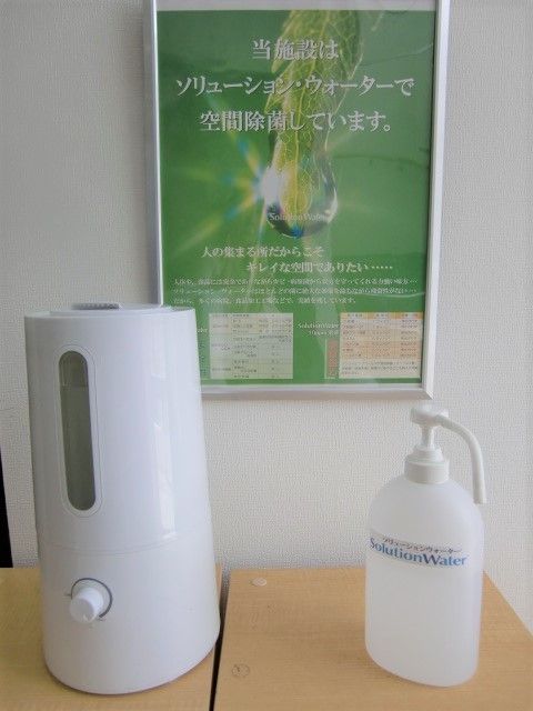白い除菌用の機械とプッシュ式の除菌液ボトルが並んで置かれ、その後ろの壁には除菌水のポスターが貼られている。