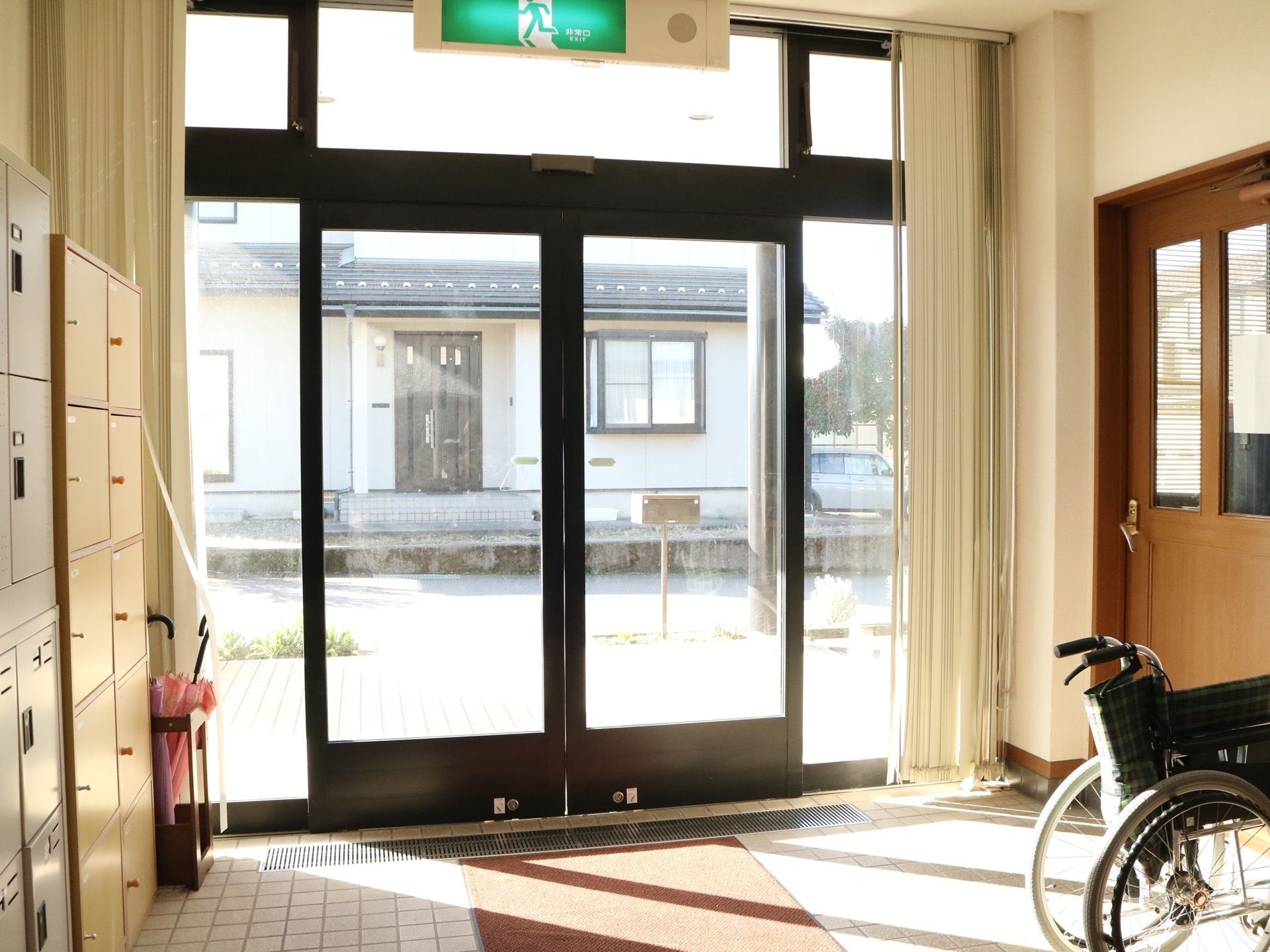 玄関入口が、黒い枠がついたガラスの両開きの自動扉である。土間に足ふきマットが敷かれていて、その近くに車椅子が折りたたんだ状態で置かれている。