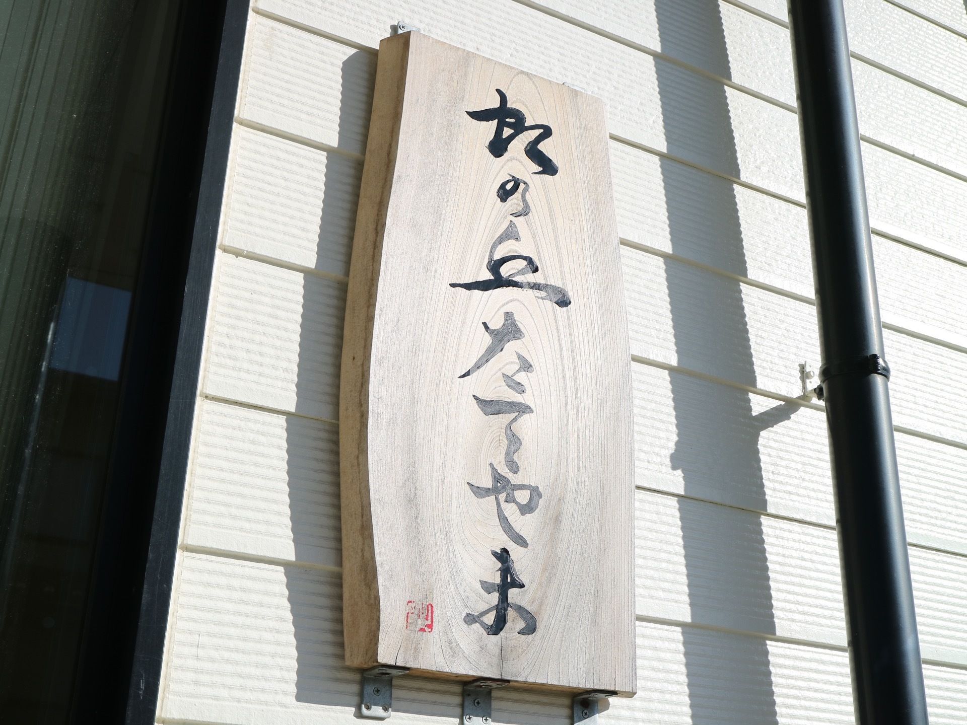 玄関扉の横の白色の壁に、縦長の木板の看板が設置されている。達筆の文字で施設名称が書かれていて、その左下に赤い落款が押されている。