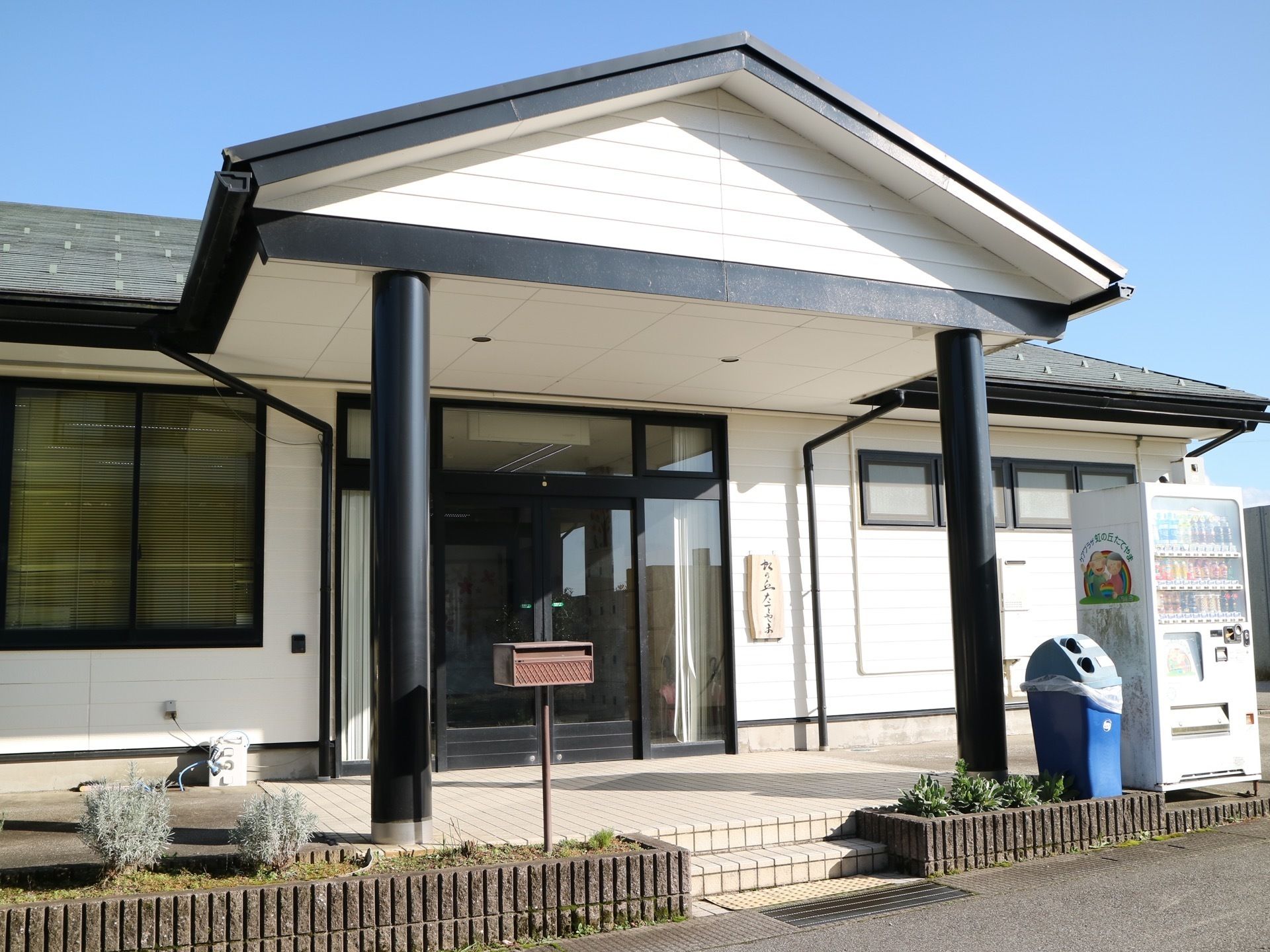 玄関前に三角屋根のひさしが張り出している。ひさしの右側に飲料の自動販売機と青色のごみ箱が置かれている。