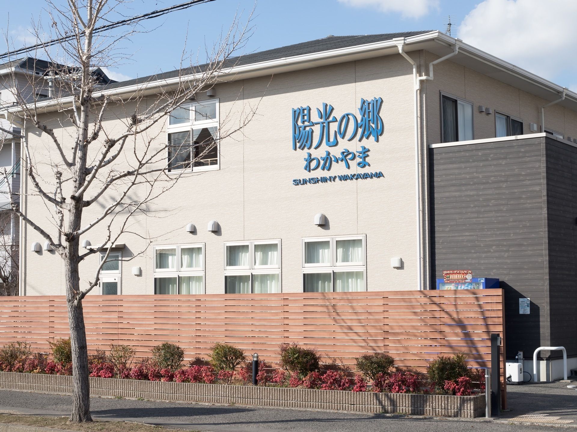 歩道沿いに木製の柵が立てられており、手前には花壇が設置されている。外壁には施設名が大きく書かれている。