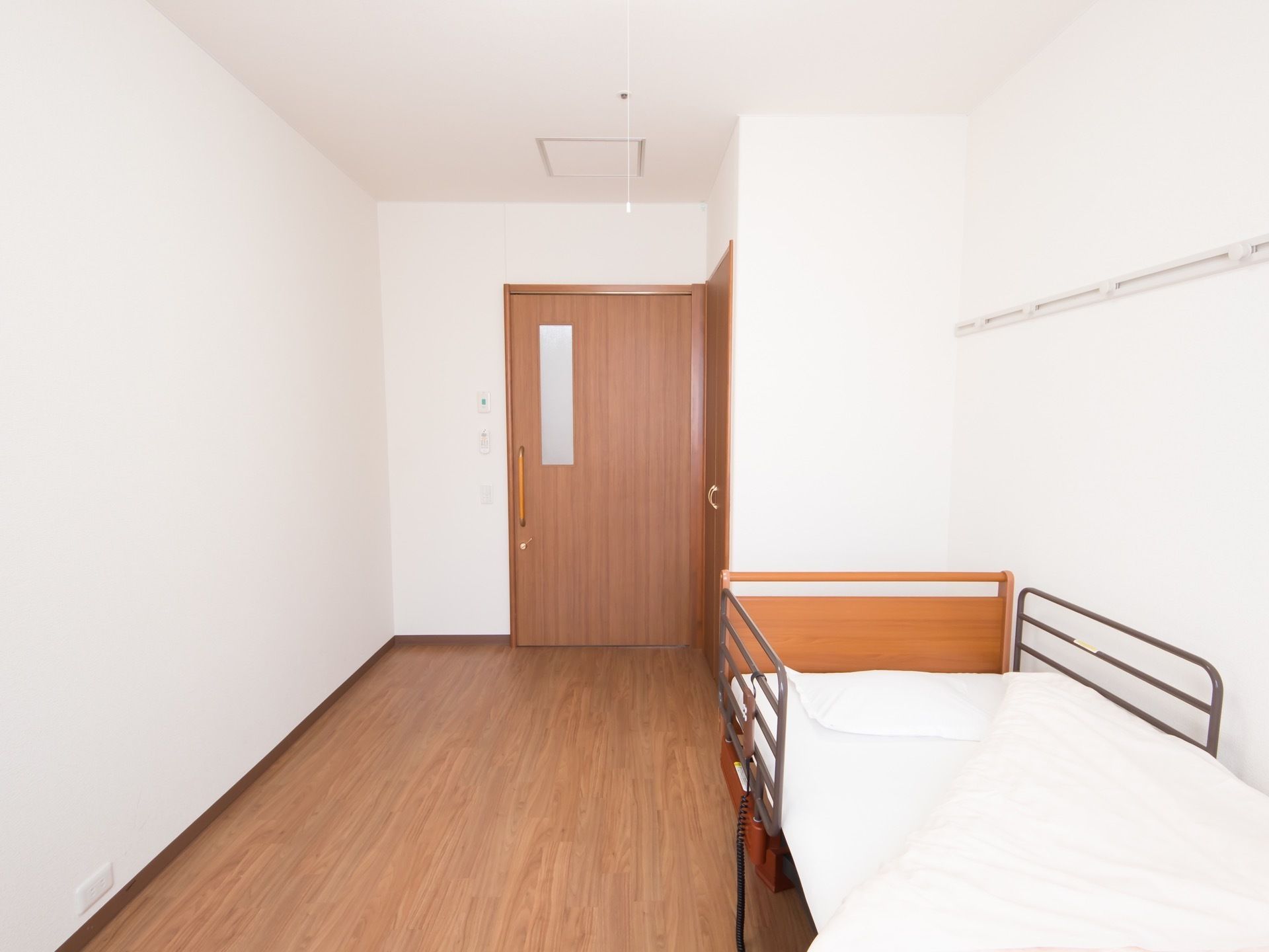 フローリングの床のワンルームの居室。シンプルなデザインの部屋で、壁沿いにはリモコンで操作可能な介護ベッドが置かれている。