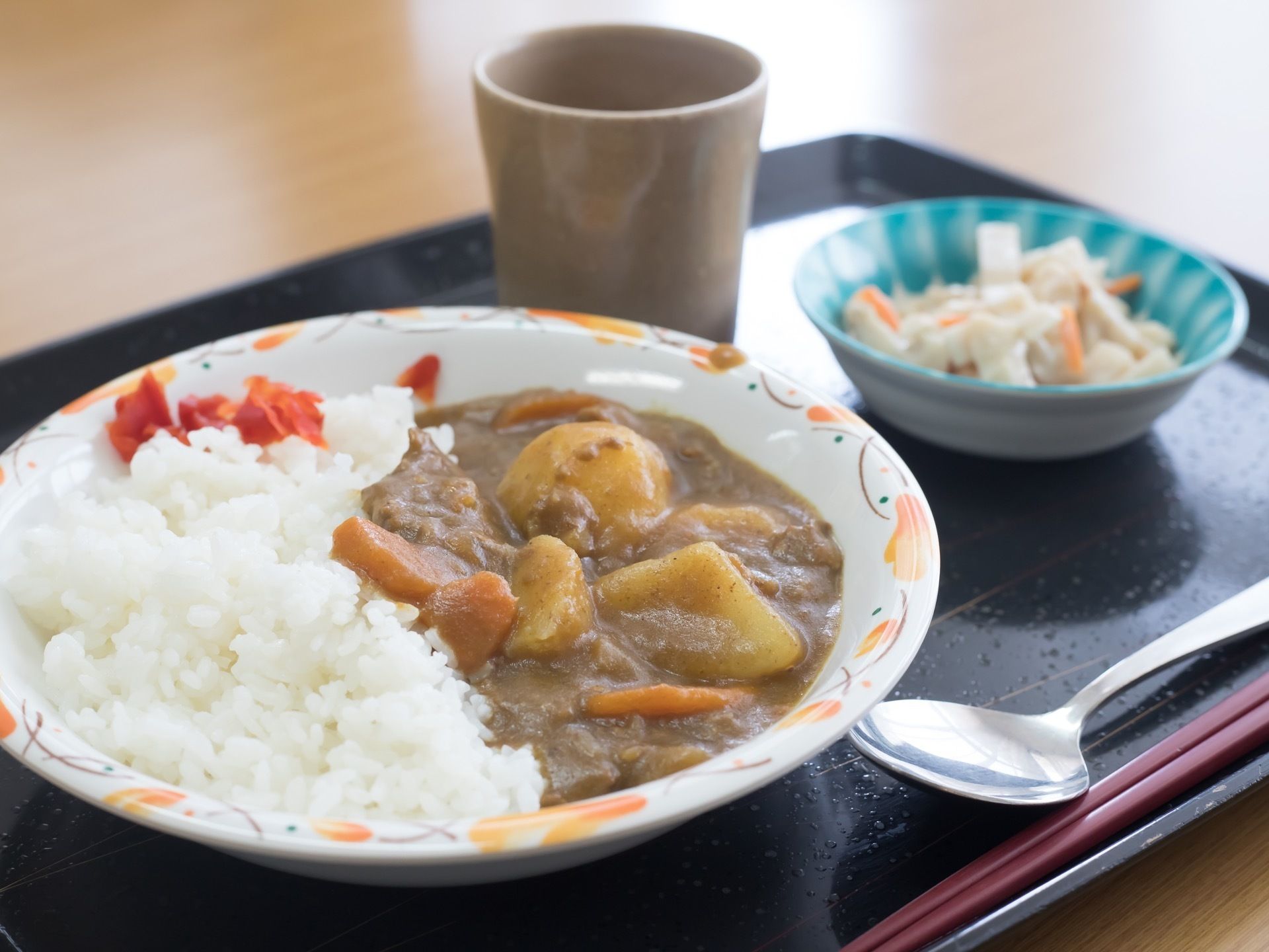 トレーの上にスプーンとお箸、カレーライス、福神漬け、サラダとお茶が用意されている。器がカラフルな柄と色である。