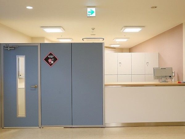 パソコンが設置されたカウンターに人はおらず、奥には白いキャビネットが置かれている。青いドアにはひし形のネームプレートが貼られている。