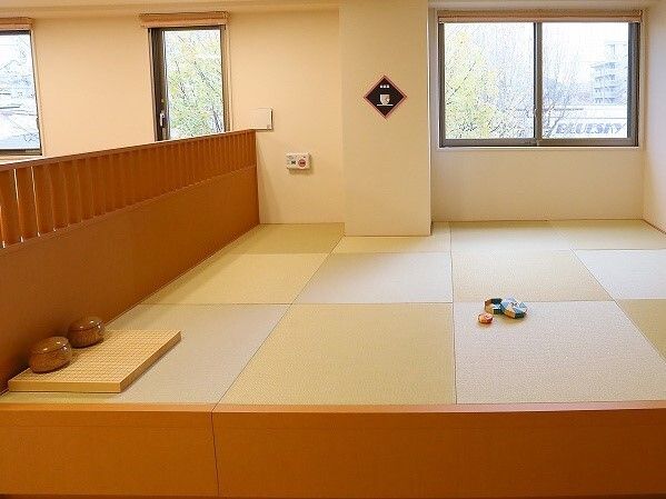 囲碁やお手玉が置かれた小上がりの畳スペース。左側には木製の柵のようなものがあり、奥の壁には赤いボタンも付いている。