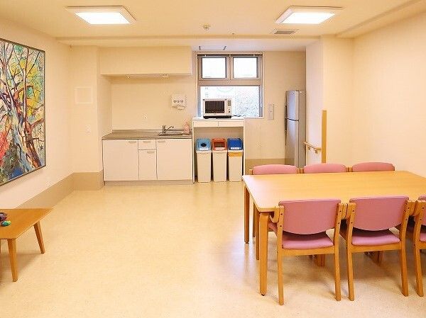冷蔵庫や電子レンジ、分別用のゴミ箱が置かれたキッチンが奥にあり、手前にはピンクの椅子がセットされたテーブルセットが置かれている。