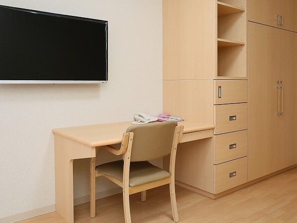 大型の収納家具や壁掛けテレビが設置されたフローリングの居室には、木製のデスクセットも置かれ、デスクの上には電話もある。