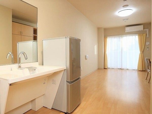 車いす対応のワイドタイプの洗面所が手前にあり、その横にはグレーの冷蔵庫が置かれ、壁にはエアコンも付いている。