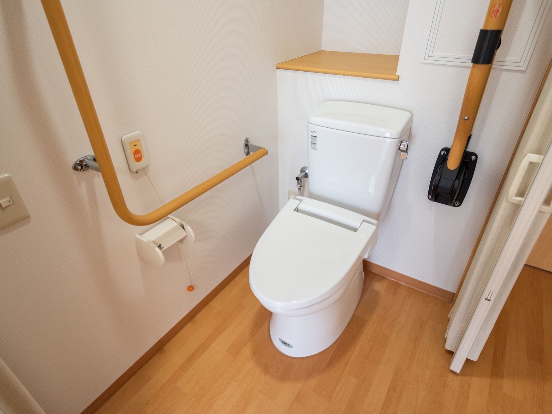 フローリングの木目調に合わせた手すりが設置されたトイレである。居室空間とはアコーディオンドアで仕切れるようになっている。