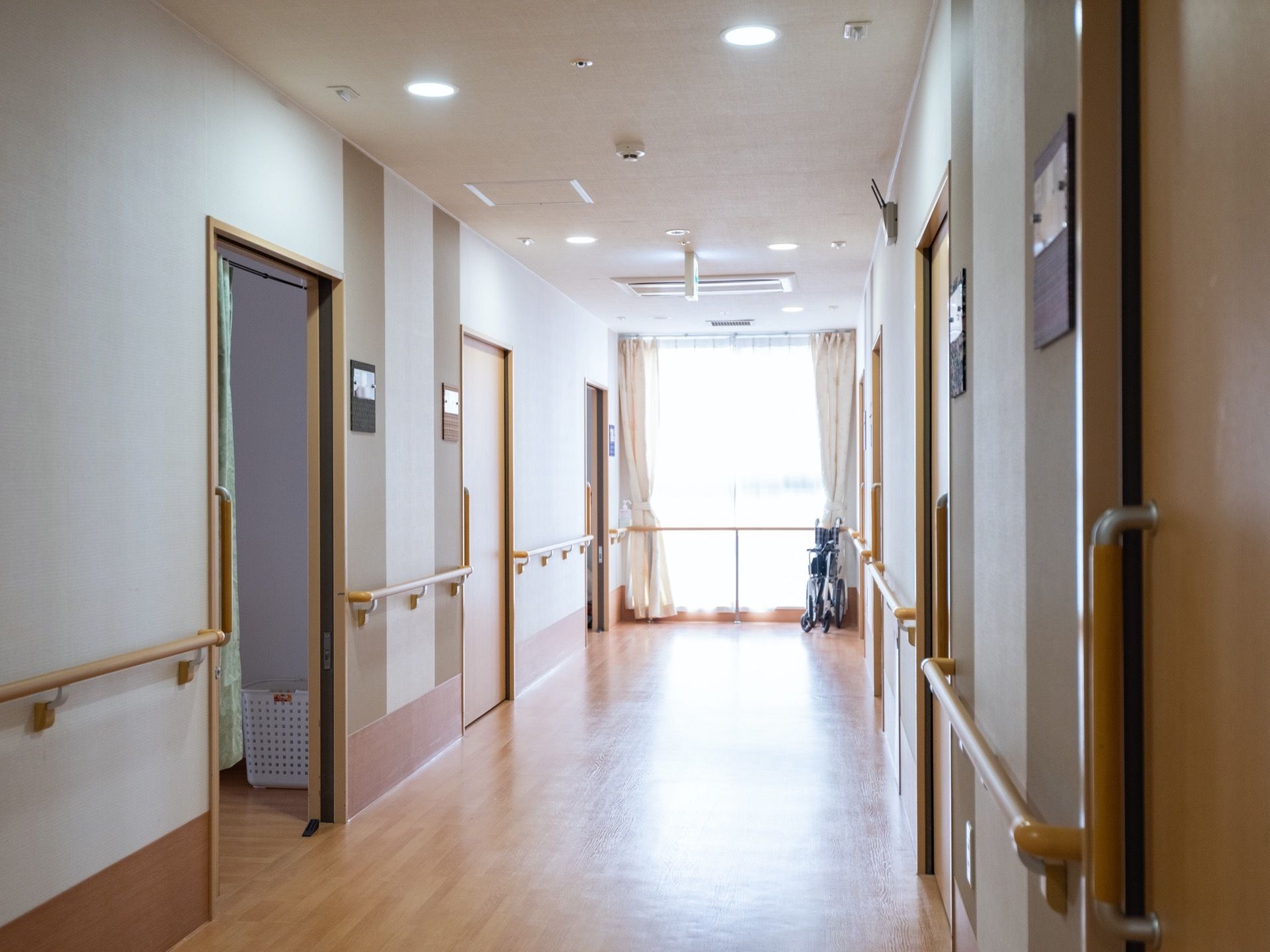 明るい光が差し込む窓が突き当りに設置された廊下である。居室の入口が並び、壁には手すりが取り付けられている。