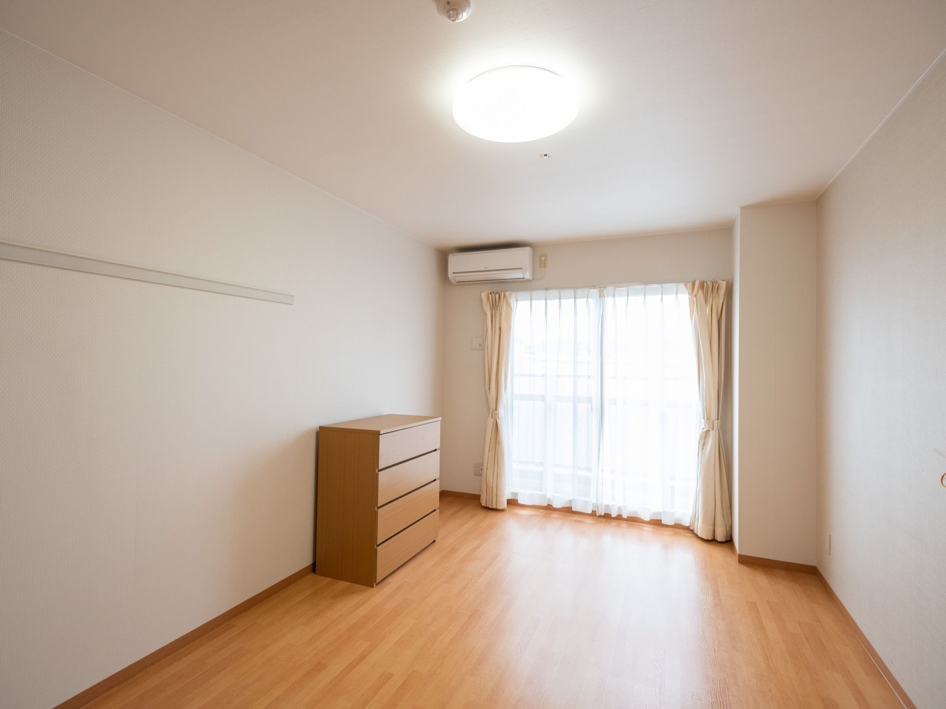 木目調のフローリング張りの床に、白い壁に囲まれた、シンプルな居室である。ピクチャーレールや照明器具、エアコンが設置されている。