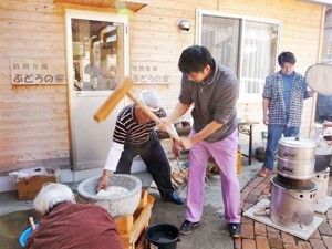 高齢の男性2名と若い男性が餅つきをしている。奥では鍋の様子を伺う若い男性がいる。
