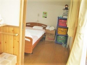 ベッドや収納家具が置かれたフローリングの居室。ベッドサイドには緊急通報装置がある。