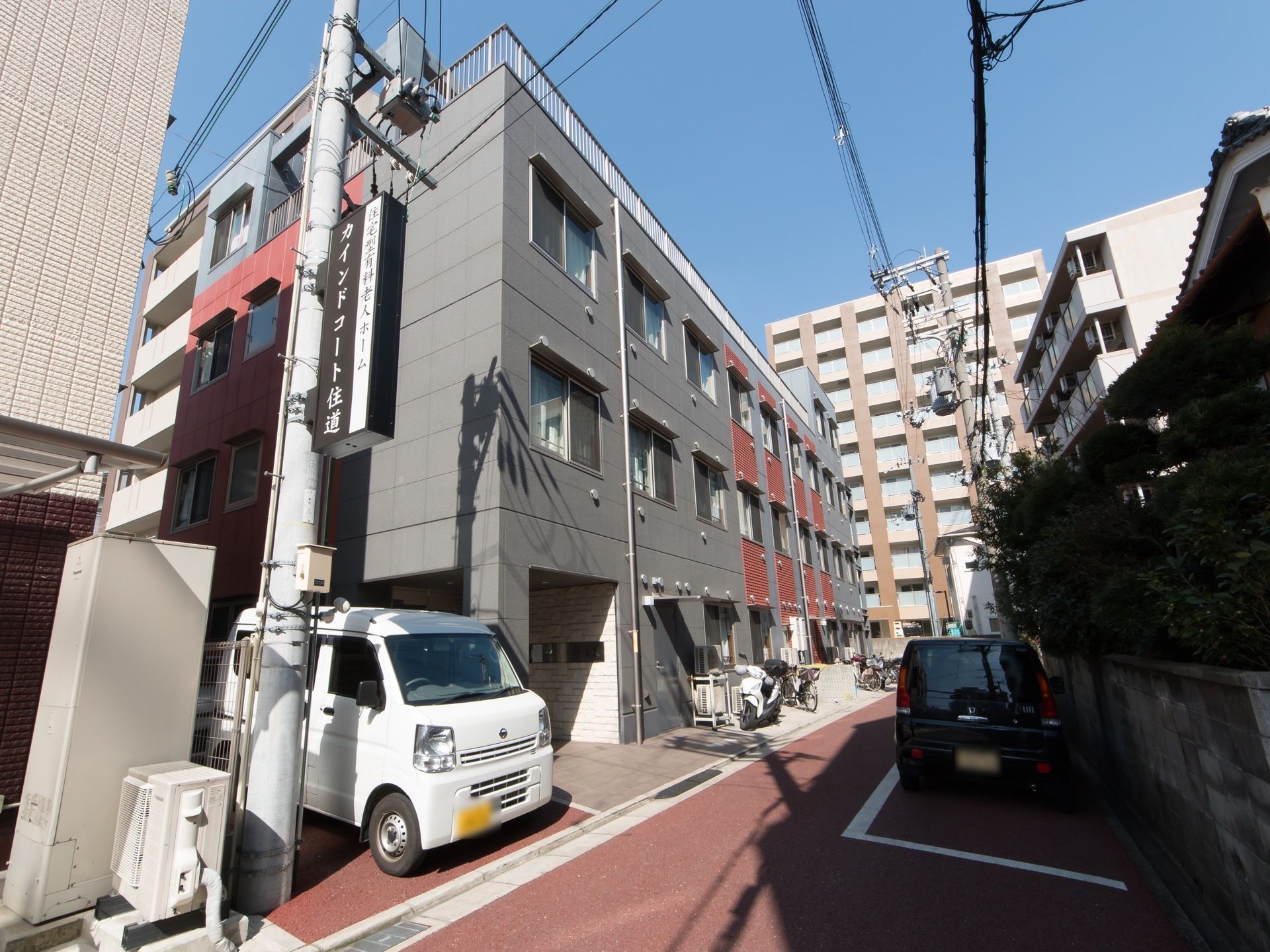 住宅街の中に建てられている３階建てのグループホーム。建物前には自転車が複数台停められており、屋上には柵が設置されている。