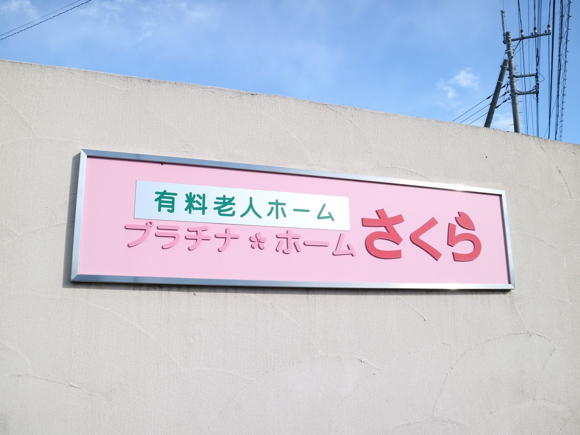 コンクリートの壁に、施設の名前が大きく書かれたピンク色の看板が貼り付けられており、周囲の景観に馴染む色合いになっている。