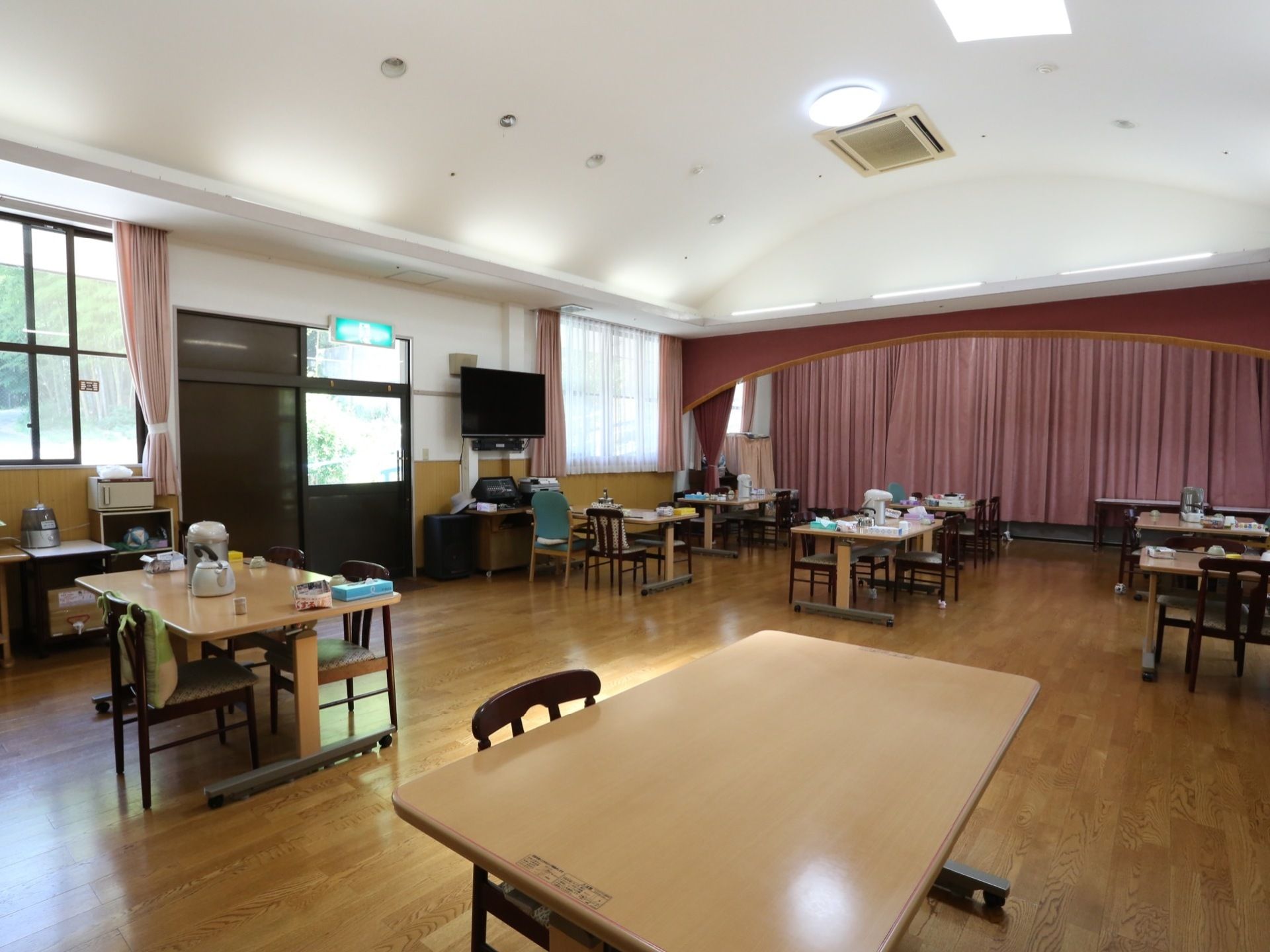 広い窓からの陽光で明るい部屋である。ダイニングテーブルと椅子が数組置かれている。中央の窓側に大型テレビがある。