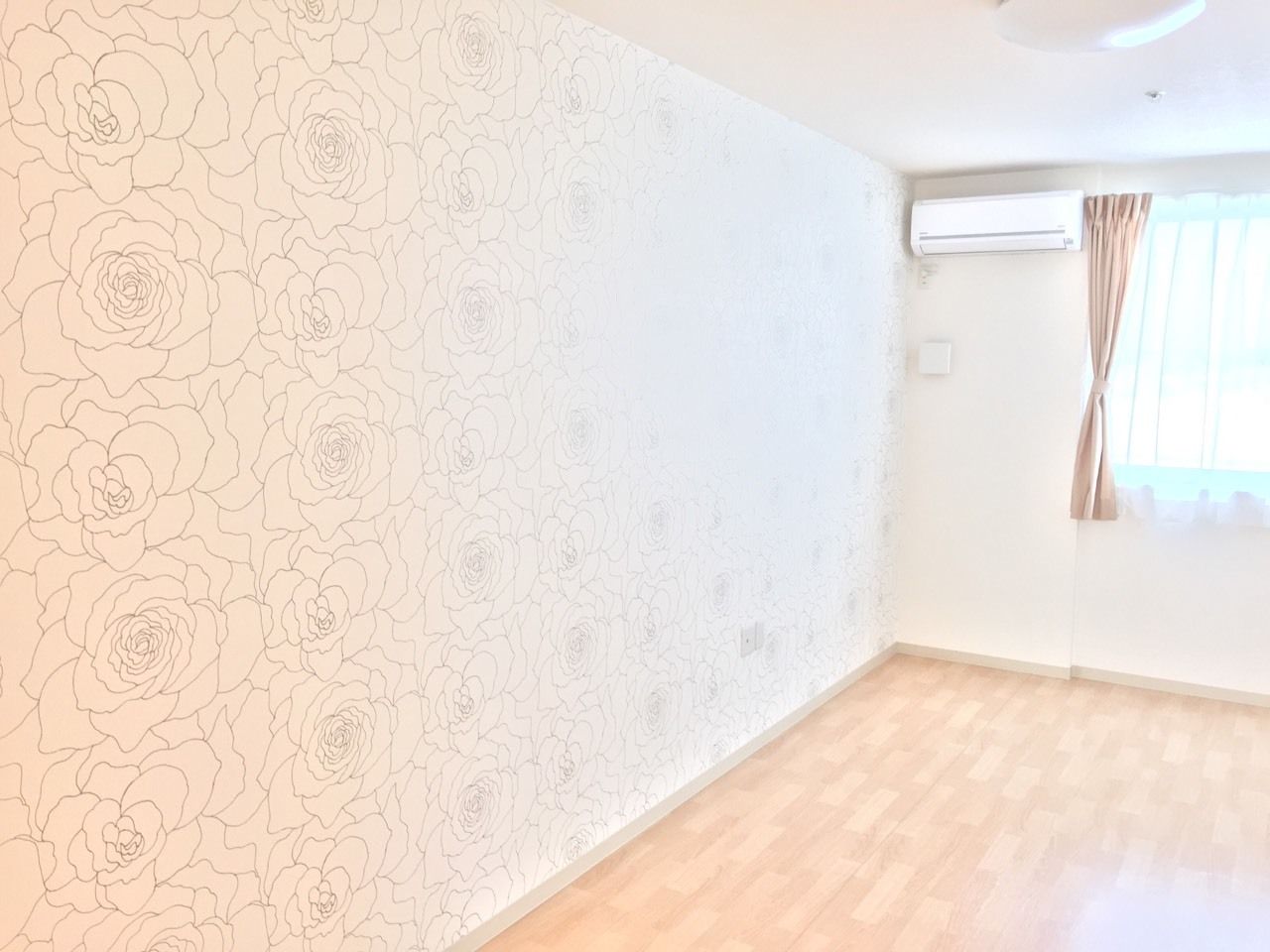 エアコンが設置されたフローリングの居室には窓が付き、壁にはバラ柄の壁紙が貼られている。