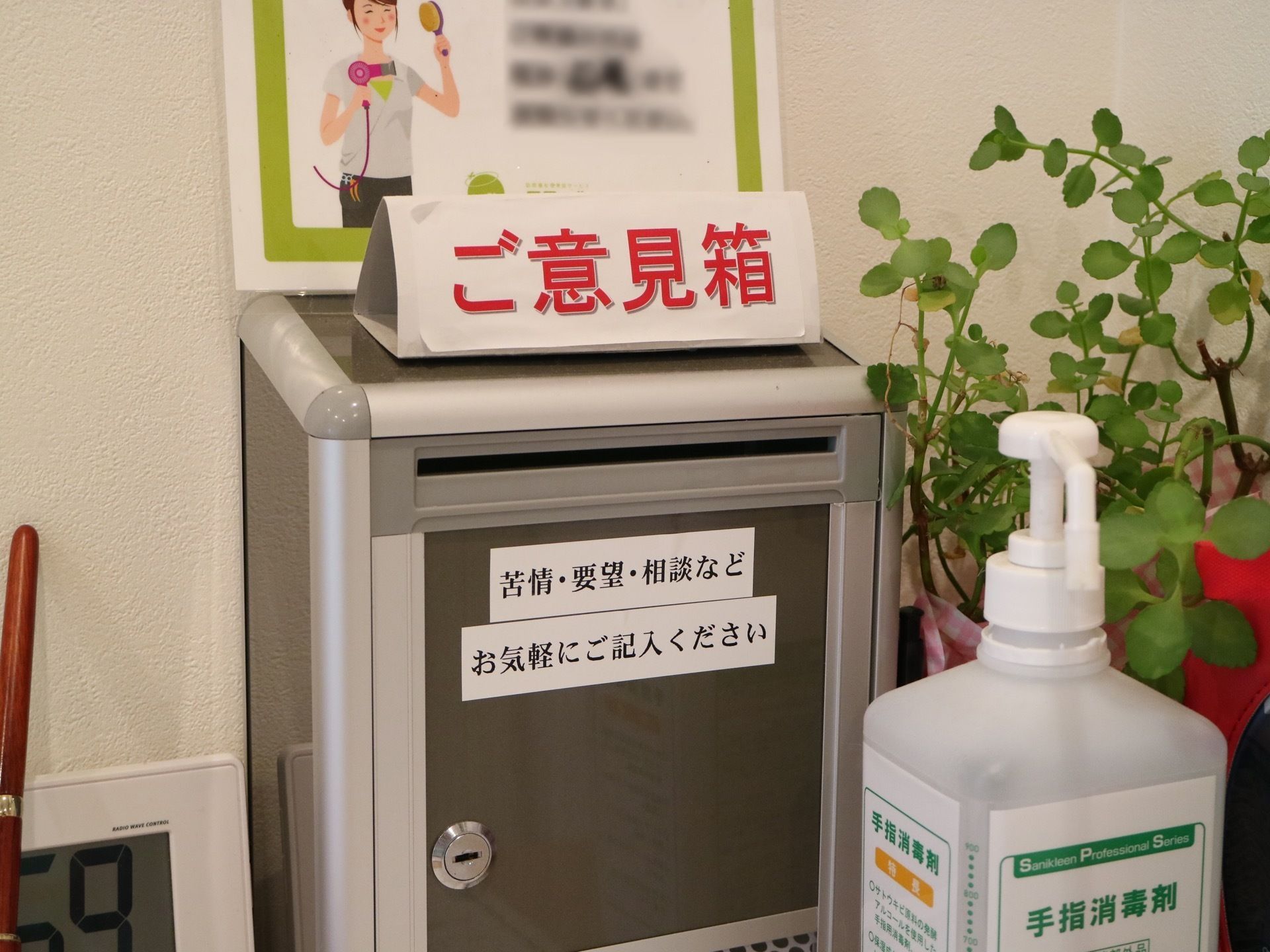ご意見箱が設置されている。鍵がかけられる。手前に手指消毒液が置かれているほか、デジタル時計も置かれている。