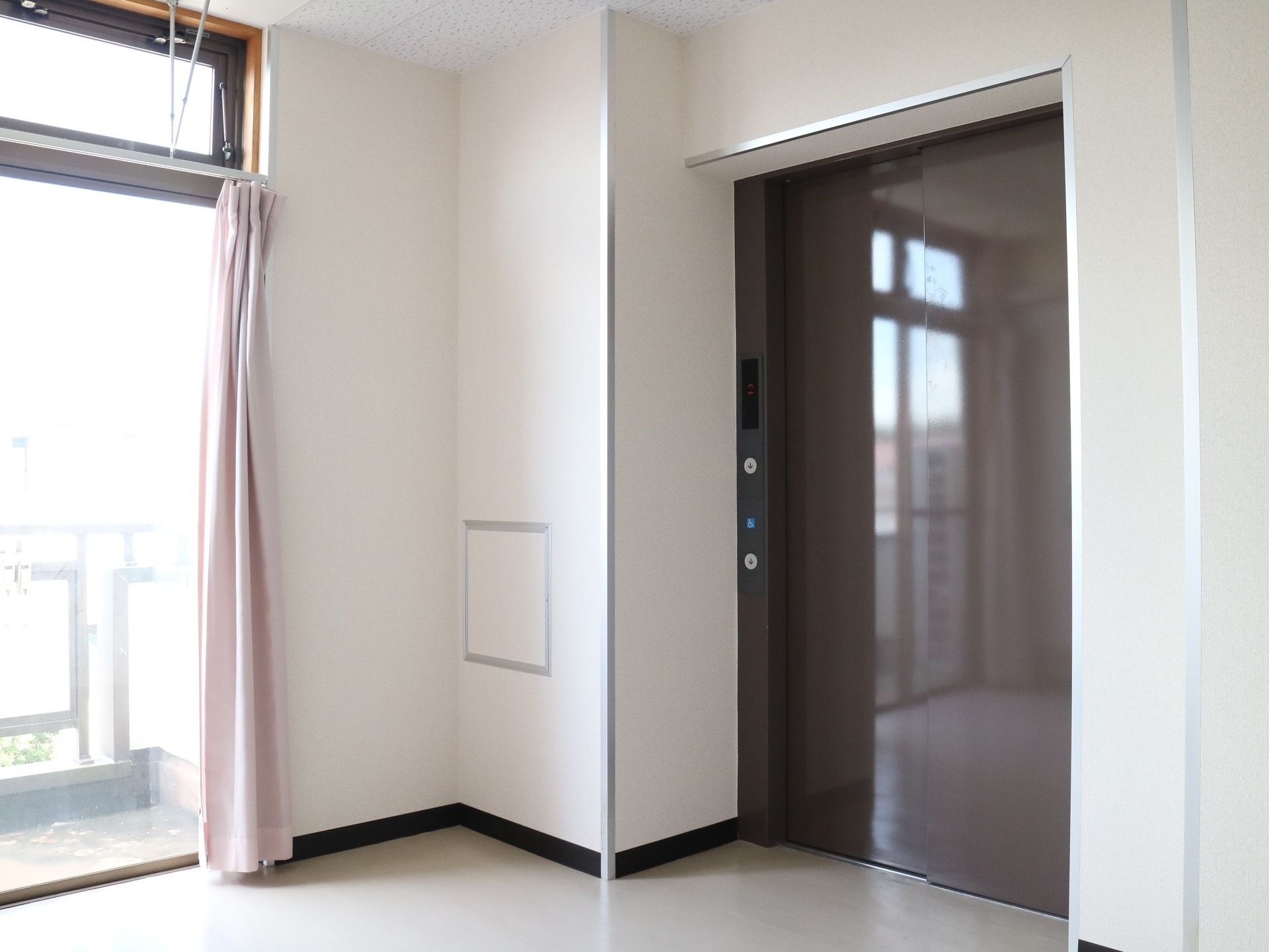 ブラウンを基調とした扉のエレベーターが１基設置されている。目の前のフロアには、ベランダへと続く大きな窓がある。
