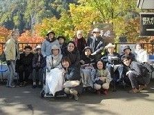 紅葉がきれいな山々をバックに集合写真を撮る入居者とスタッフ。車いすを利用している入居者もいる。