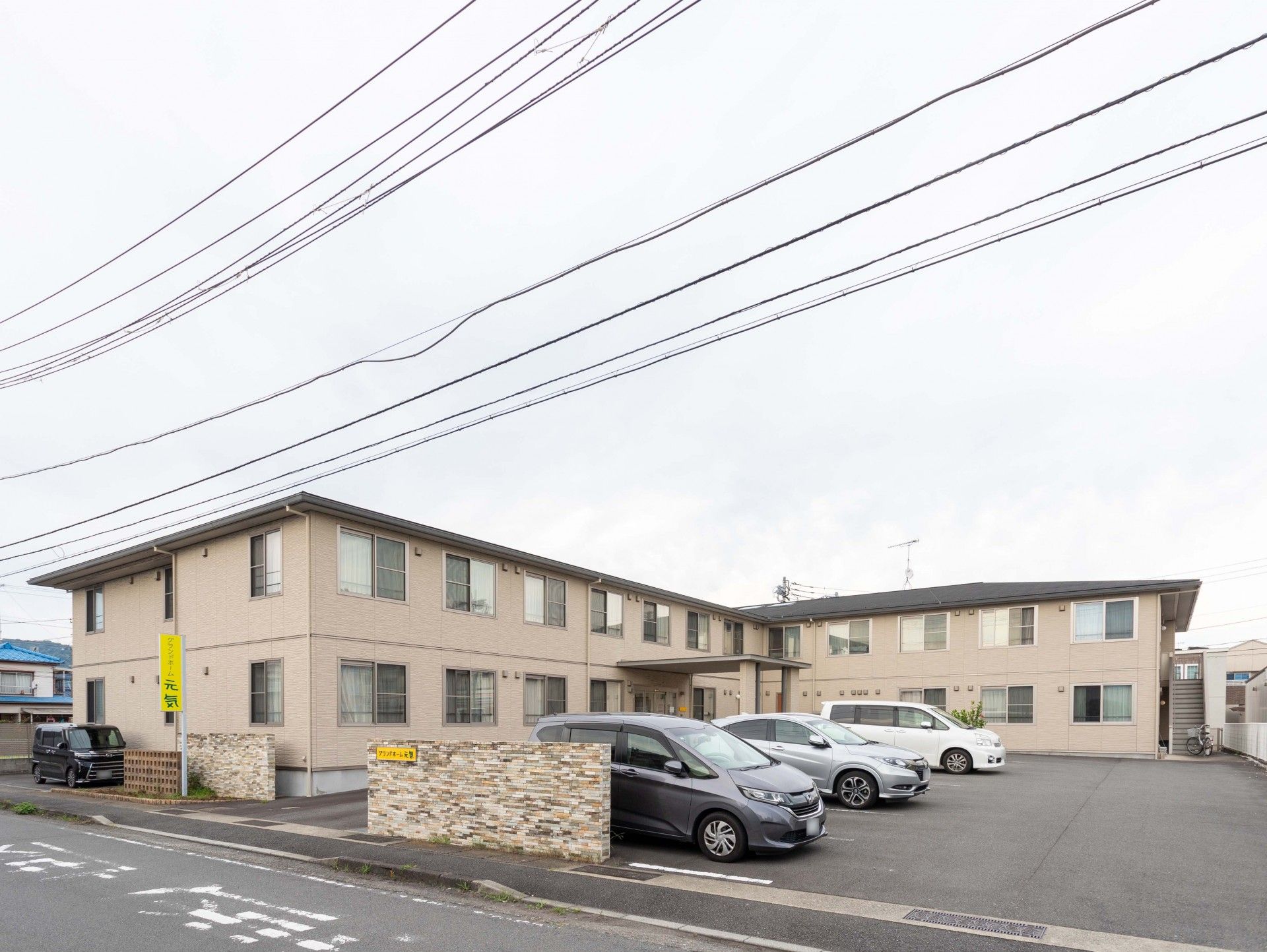 広い駐車場があり、２階建ての建物は日当たりのいい場所にあり、ベージュの外壁でエル字型。