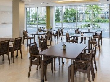 一輪挿しが飾られた4人掛けのテーブルセットが複数置かれた食堂。大きな窓の外には草木が植えられた駐車場がある。