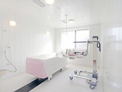白とピンクの機械浴が設置された浴室には大きな窓があり、ストレッチャータイプのリフトが浴槽タイプの機械の横にある。