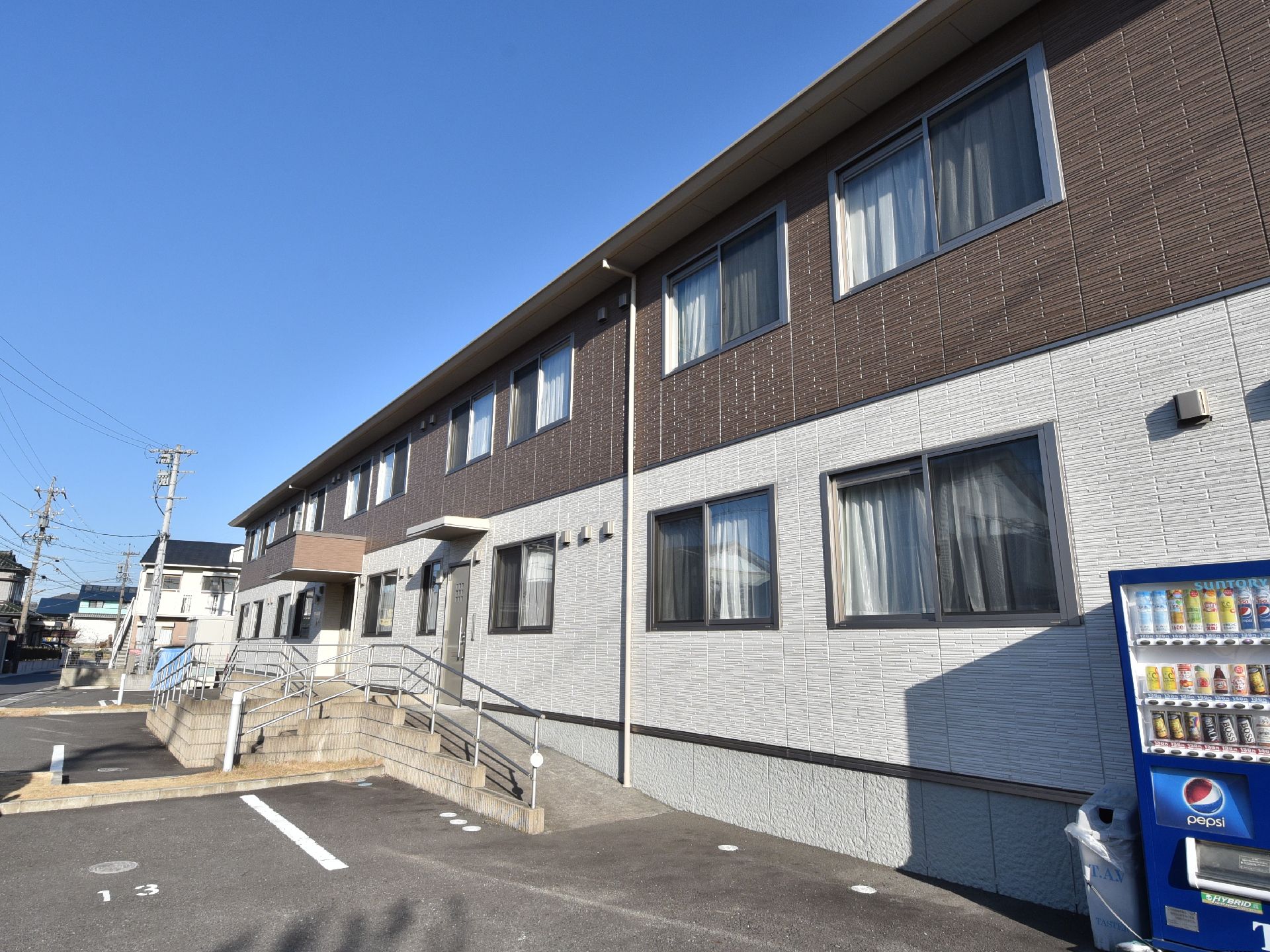 白とダークブラウンの２階建ての建物は玄関に屋根があり、手すりが付いたスロープが設置されている。