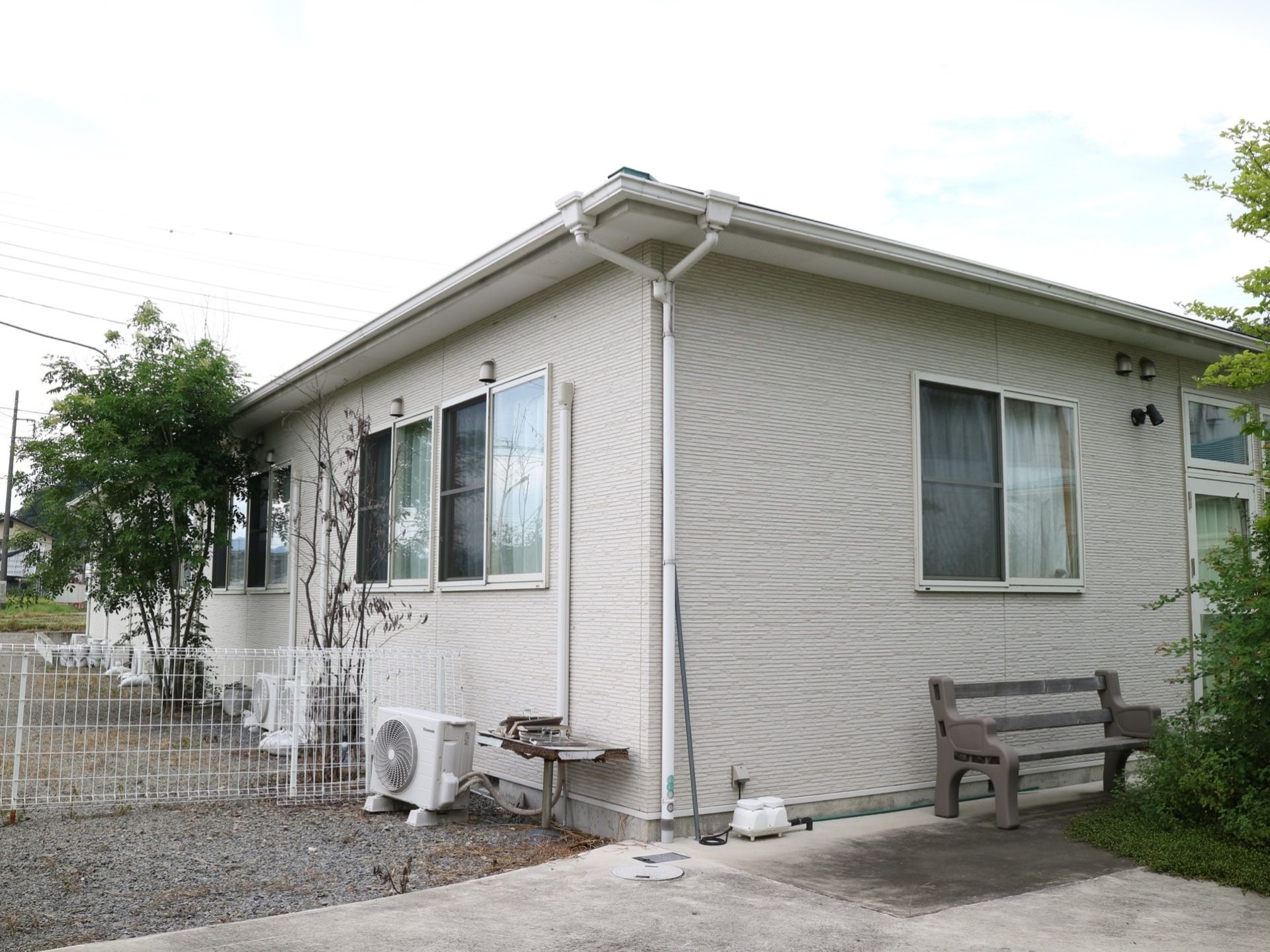 薄いグレーの外壁の平屋建ての建物である。周囲には、白いフェンスがあり、壁際には木製のベンチが置かれている。