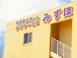 山吹色の外壁の建物には屋外階段が付いている。外壁にはホーム名と併設事業所名が書かれている。