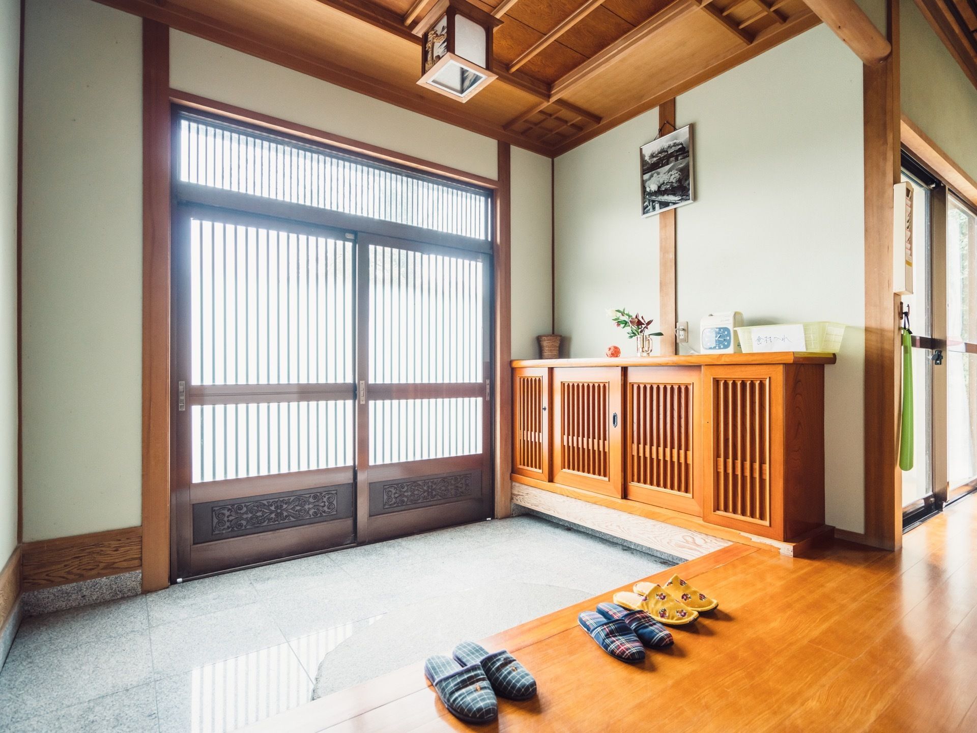 和風建築の格子状の引き戸の玄関がある。下駄箱が置かれ、花や物が置かれている。玄関を上がったところにスリッパが並んでいる。