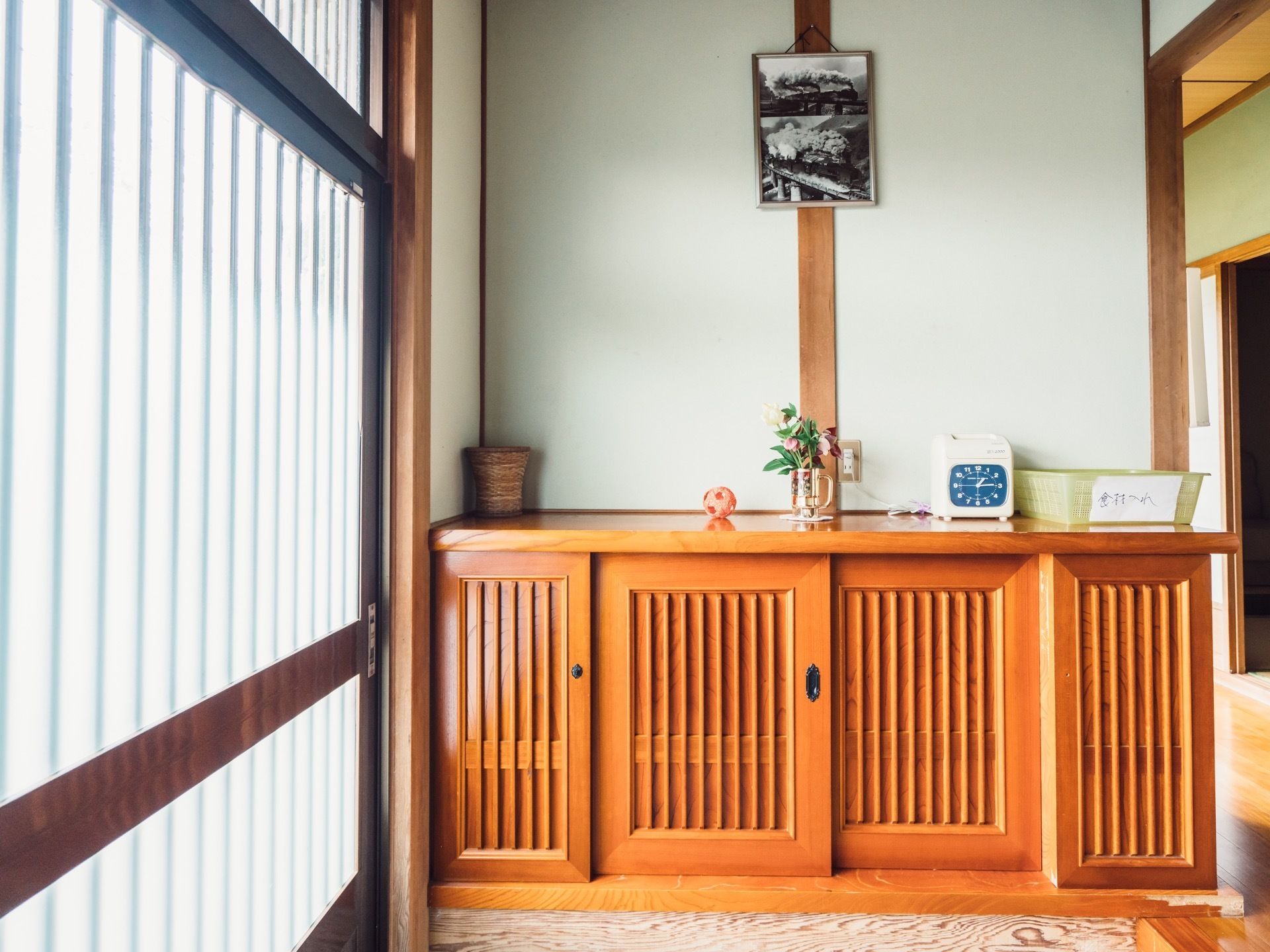格子状の引き戸のある玄関。どっしりとした木の下駄箱が置かれている。花や物が置かれ、壁には古いモノクロ写真が飾られている。