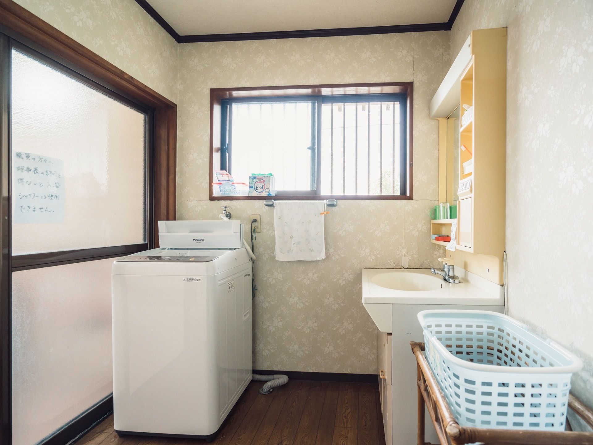 浴室へと続く脱衣所に、全自動洗濯機が置かれている。窓がついており、棚と鏡のついた洗面台と脱衣かごがある。