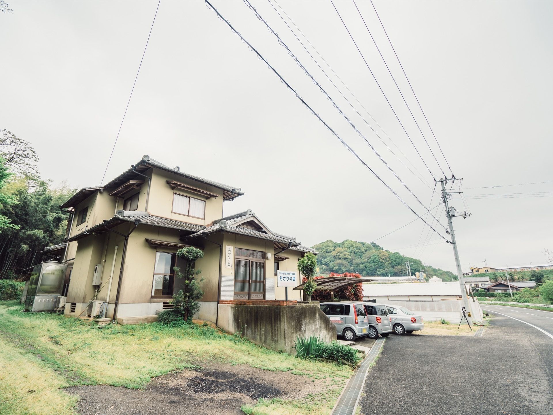 道路に面した場所にある、２階建ての日本建築の建物。屋根は瓦造りになっている。周囲は緑が生い茂り、側には車が数台停まっている駐車場がある。
