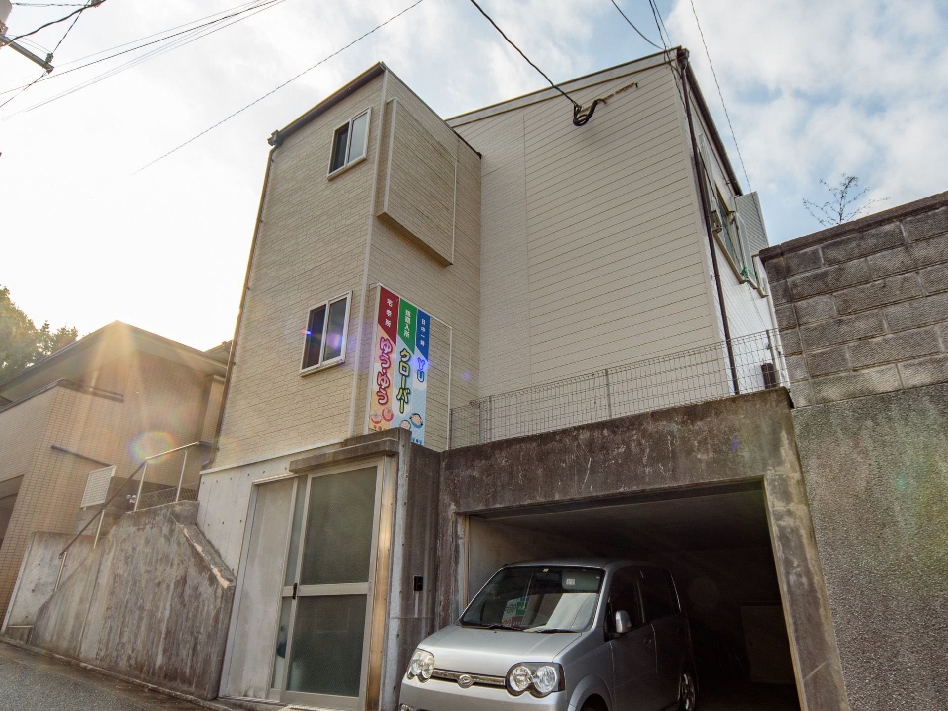 1階部分に駐車場があり、自動車が停まっている。2階に当たるところが建物全体の1階があり、道路から建物まで階段が設置されている。