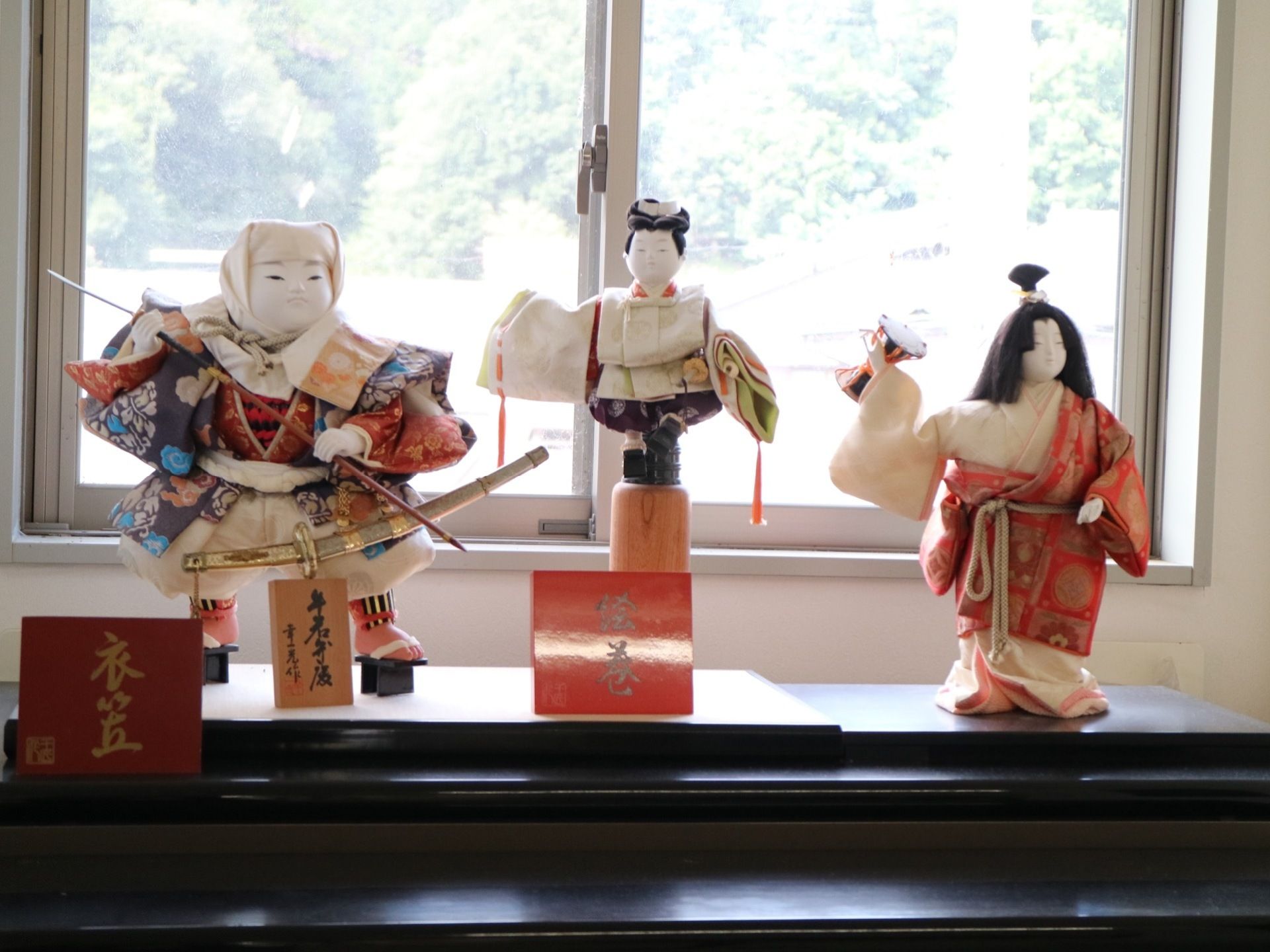窓際の棚の上に日本人形が３体並べられている。人形が現わしている人物や作品名が表示された小さな板が添えられている。