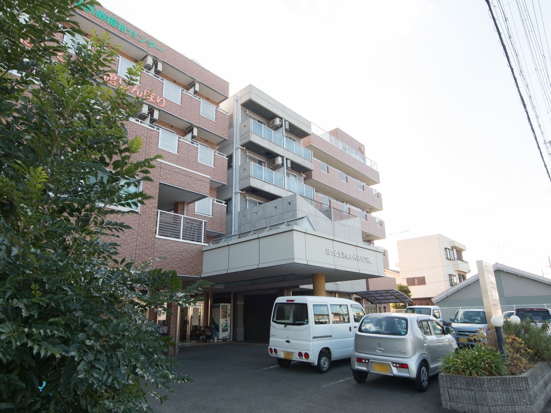 ５階建ての施設である。建物の外壁の上の方に大きな文字で施設名称が表示されている。玄関前に車が何台も停まっている。